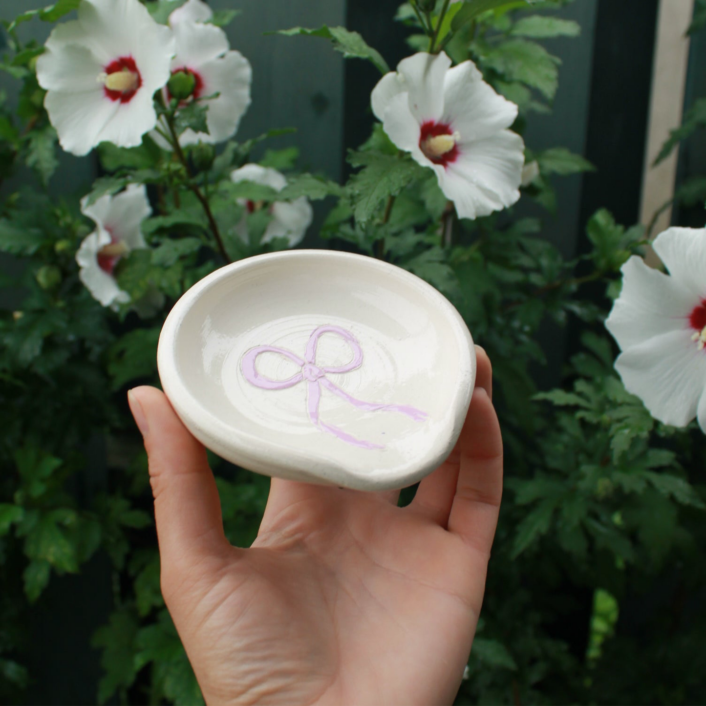 hand thrown off white spoon rest decorated with a lavender purple bow in the centre. held in hand against a dark green foliage background, with white and pink hibiscus flowers peeking out.