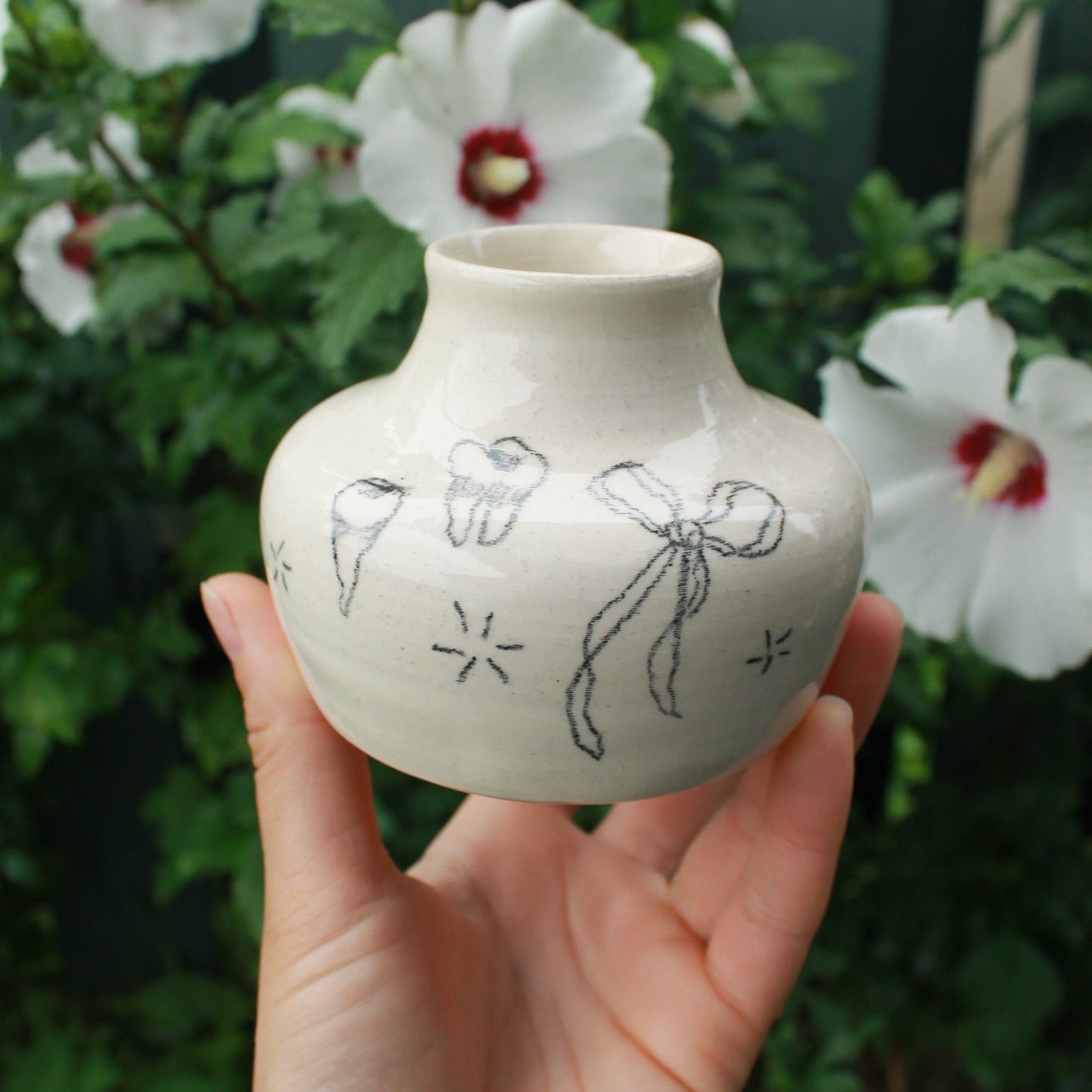 Hand holding a small ceramic vase with a flowy bow and teeth designs drawn on it. In the background are some white hardy hibiscus flowers tucked into some dark green foliage.