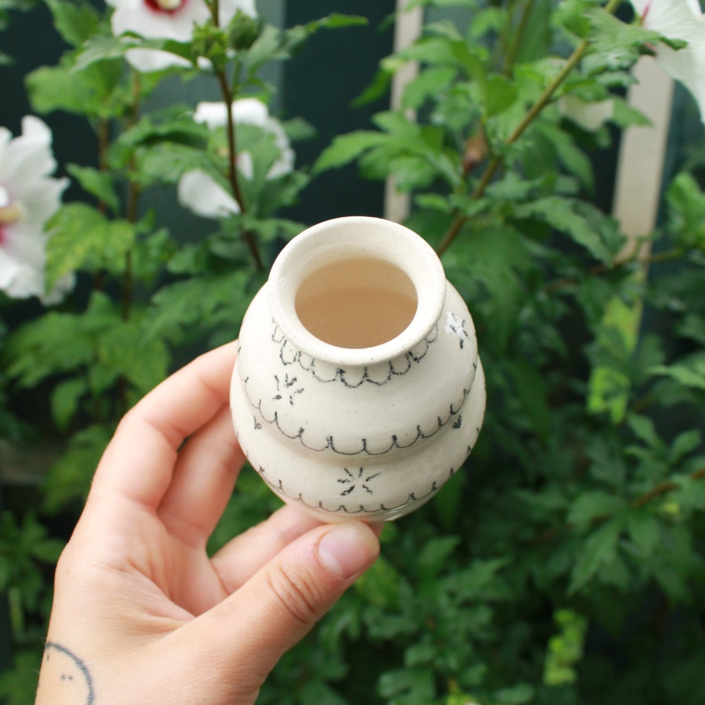 handthrown bubbly bud vase made from white clay, decorated with black pencil scalloped lines and tiny starbursts and hearts. held in hand against a dark foliage background with white hibiscus flowers.