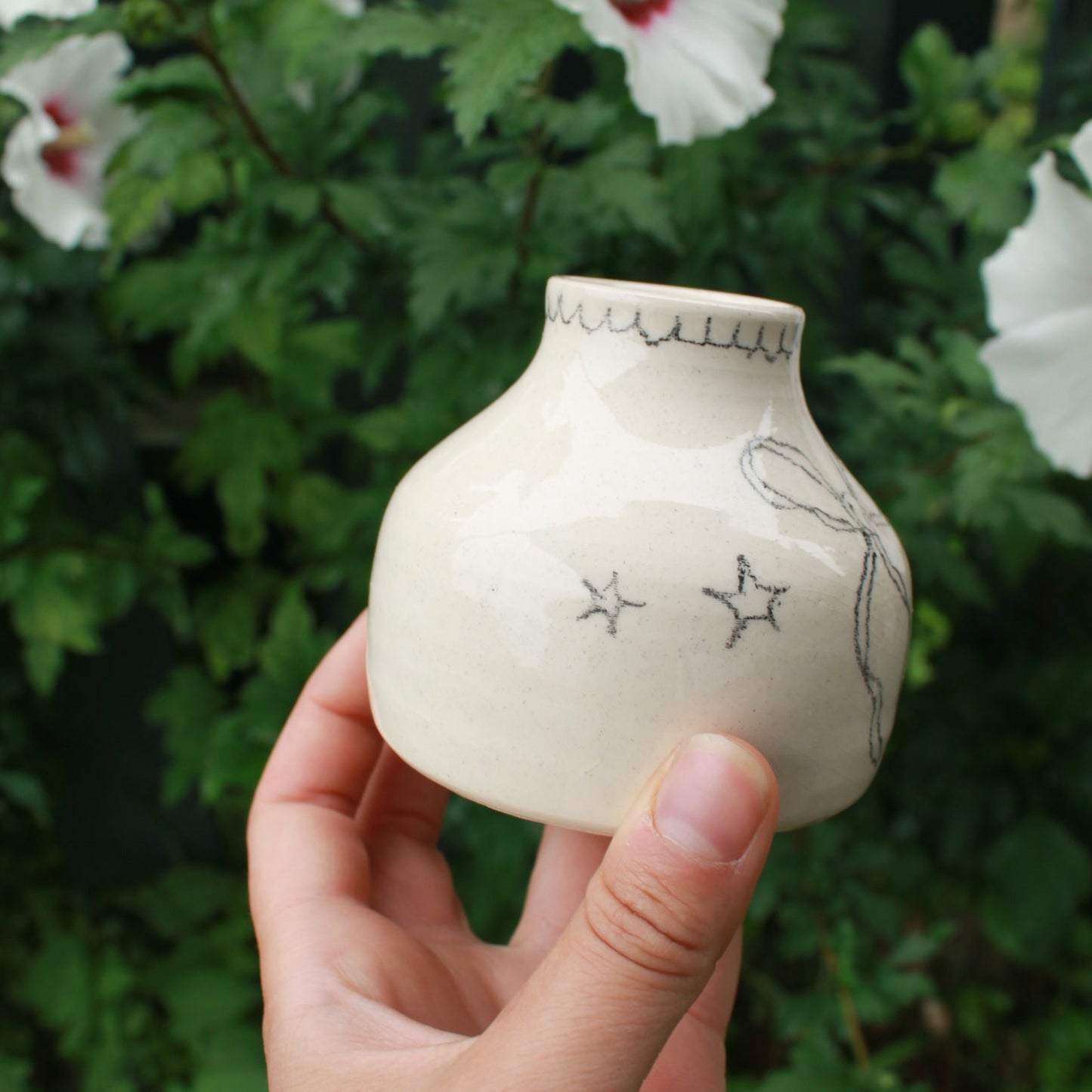 hand holding a small off white ceramic vase with star and bow design, and a scalloped edge drawn around the opening. background is dark green foliage with pink and white hibiscus flowers peeking out.