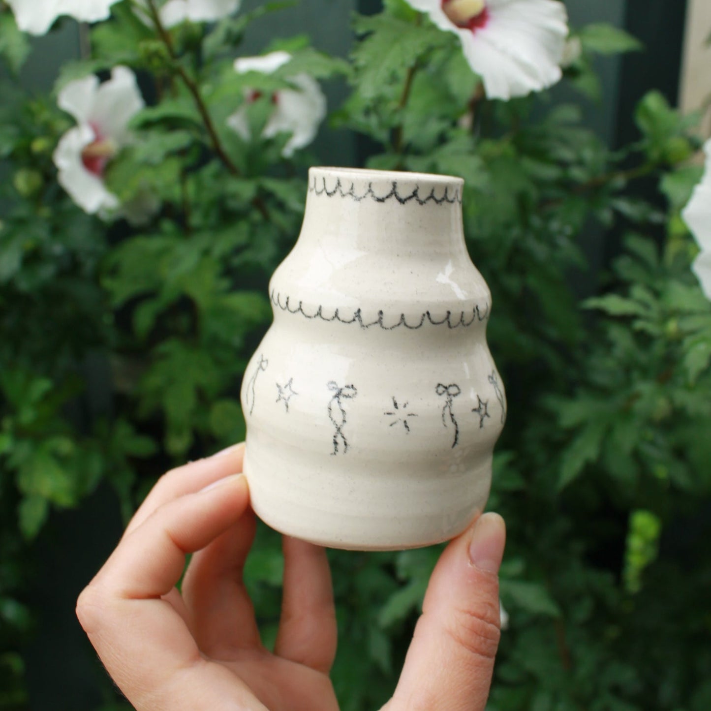 handthrown bubble vase with long neck, decorated with bows, starbursts and scalloped lines. held in hand against a dark foliage background and hibiscus flowers.