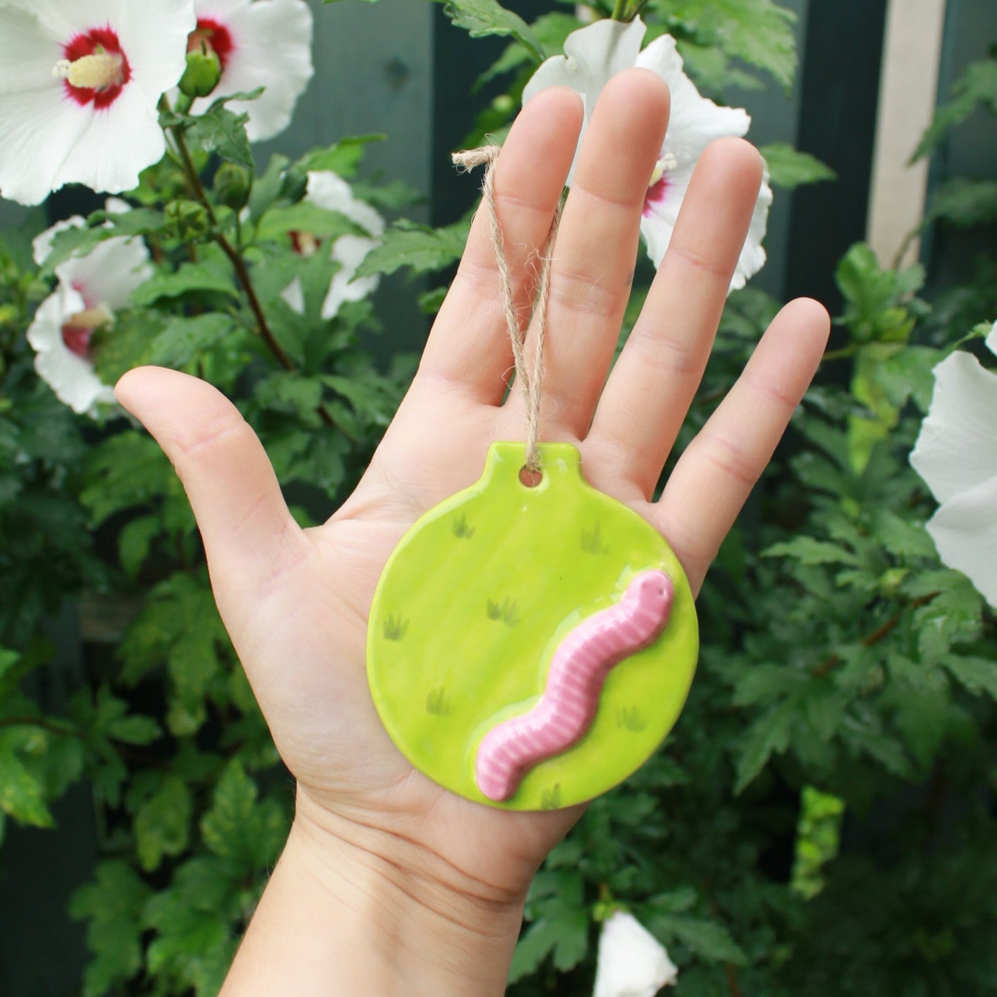 flat circular ornaments with pink worms appliqued to them. The design is a green meadow. held in hand against a dark green foliage background, with white and pink hibiscus flowers peeking through.