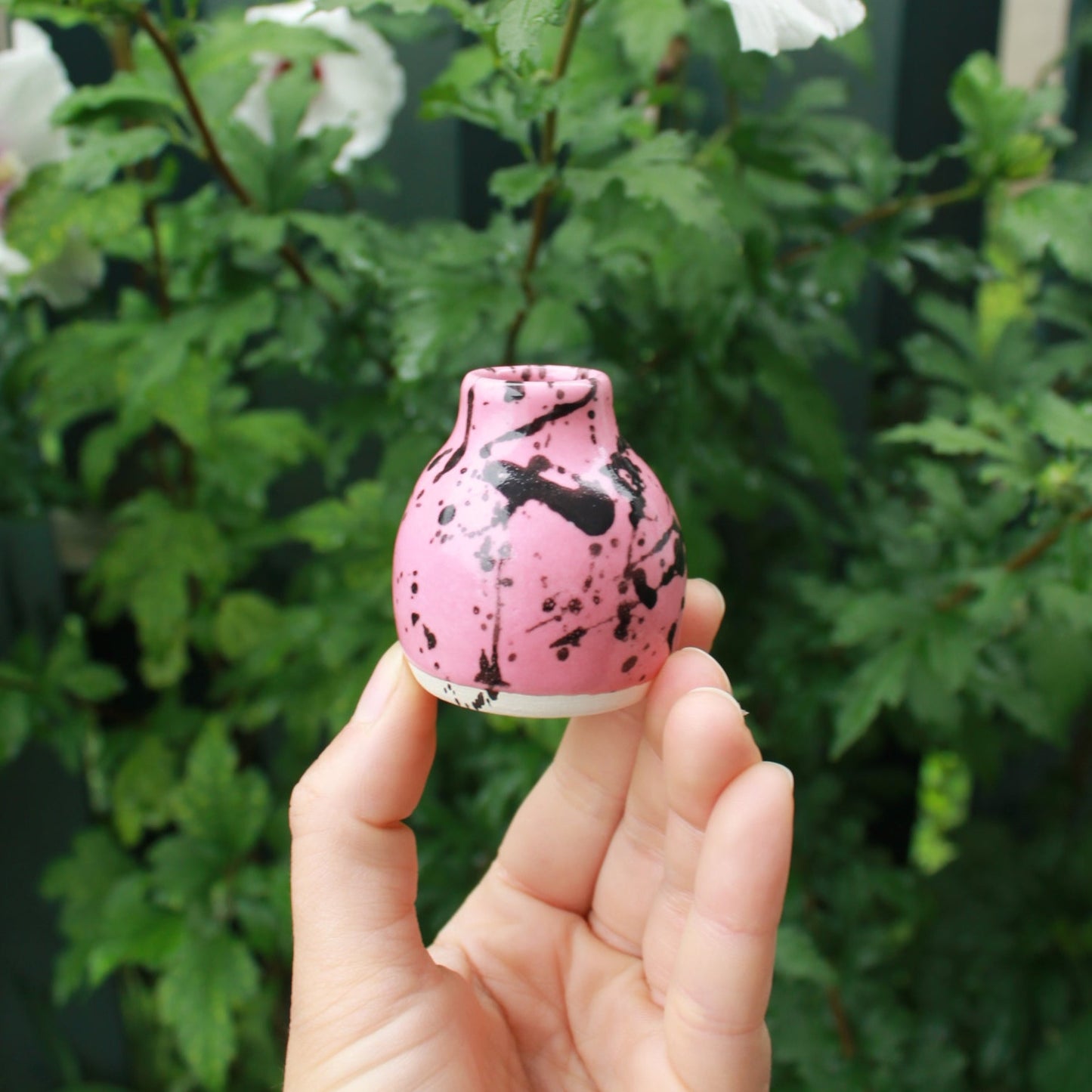 tiny pink bud vase with a shoulder and small neck, decorated with pink glaze and black splatter. held in hand against a dark foliage background with hibiscus flowers.
