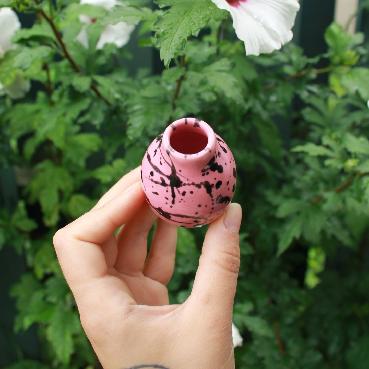 tiny pink bud vase with a shoulder and small neck, decorated with pink glaze and black splatter. held in hand against a dark foliage background with hibiscus flowers.