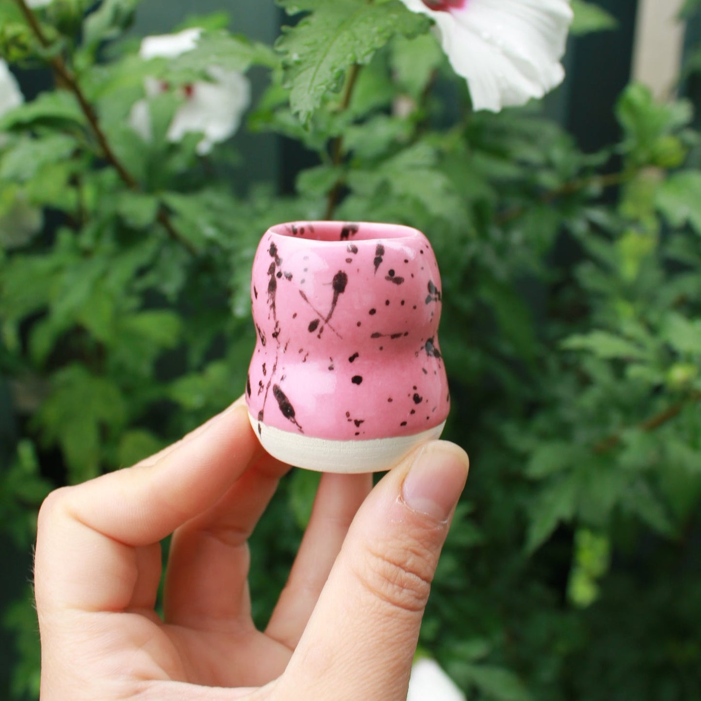 micro bubble bud vase decorated with pink glaze and black splatter. held in hand against a dark foliage background with hibiscus flowers.