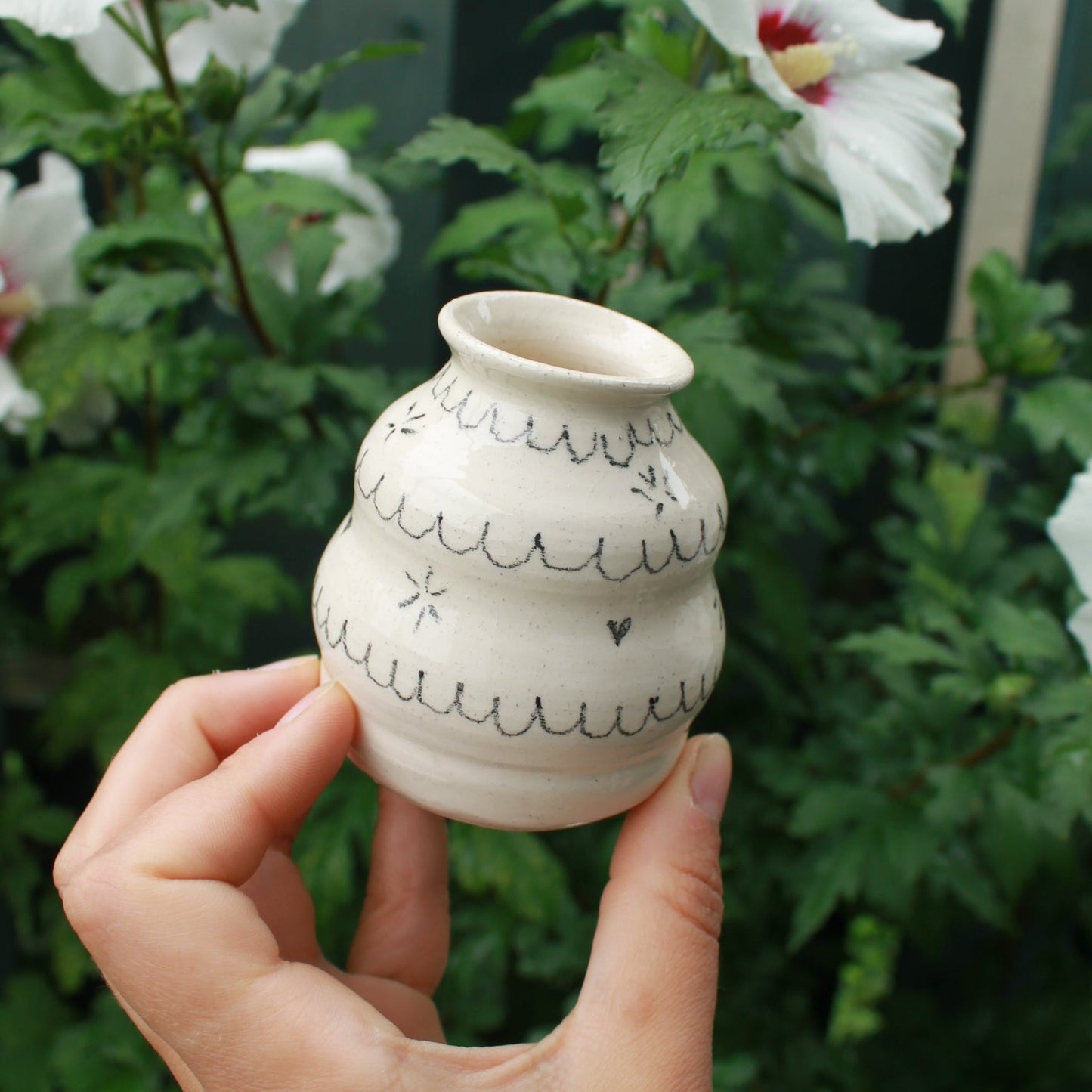 handthrown bubbly bud vase made from white clay, decorated with black pencil scalloped lines and tiny starbursts and hearts. held in hand against a dark foliage background with white hibiscus flowers.