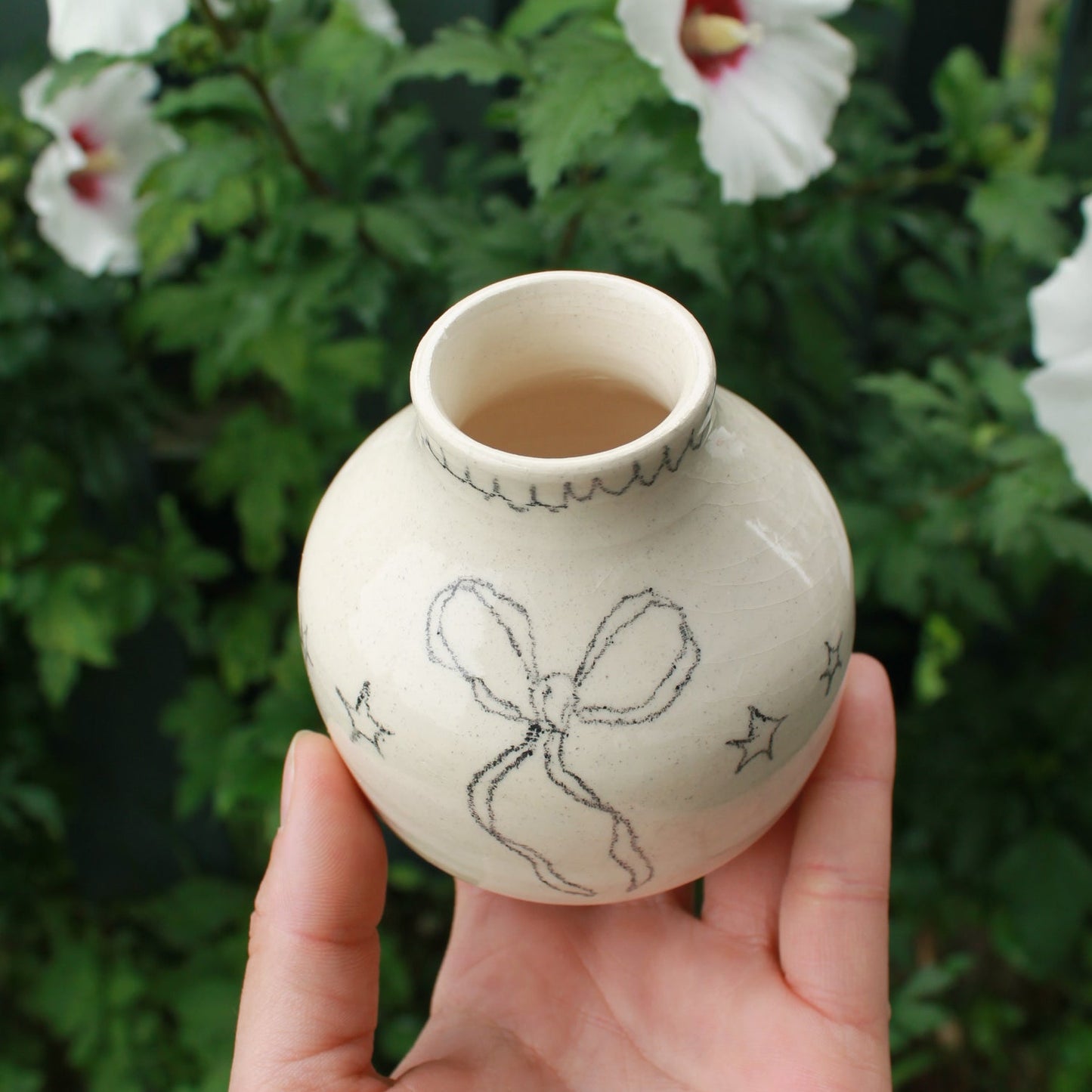 hand holding a small off white ceramic vase with star and bow design, and a scalloped edge drawn around the opening. background is dark green foliage with pink and white hibiscus flowers peeking out.