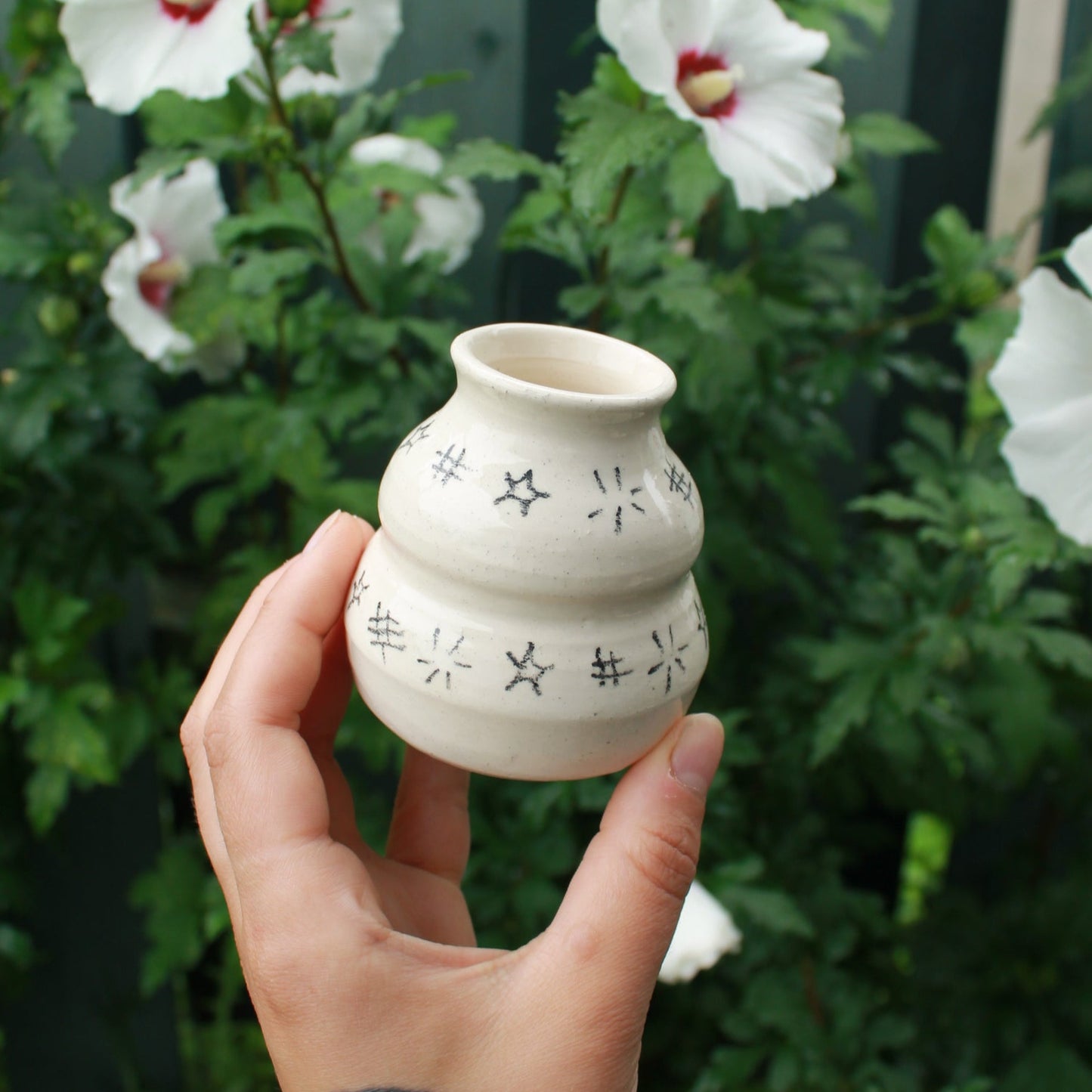 small handthrown bubble vase decorated with hatching and starbursts in black pencil. held in hand against a dark foliage with hibiscus flowers.