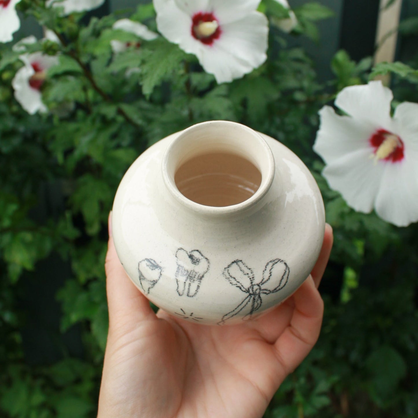 Hand holding a small ceramic vase with a flowy bow and teeth designs drawn on it. In the background are some white hardy hibiscus flowers tucked into some dark green foliage.
