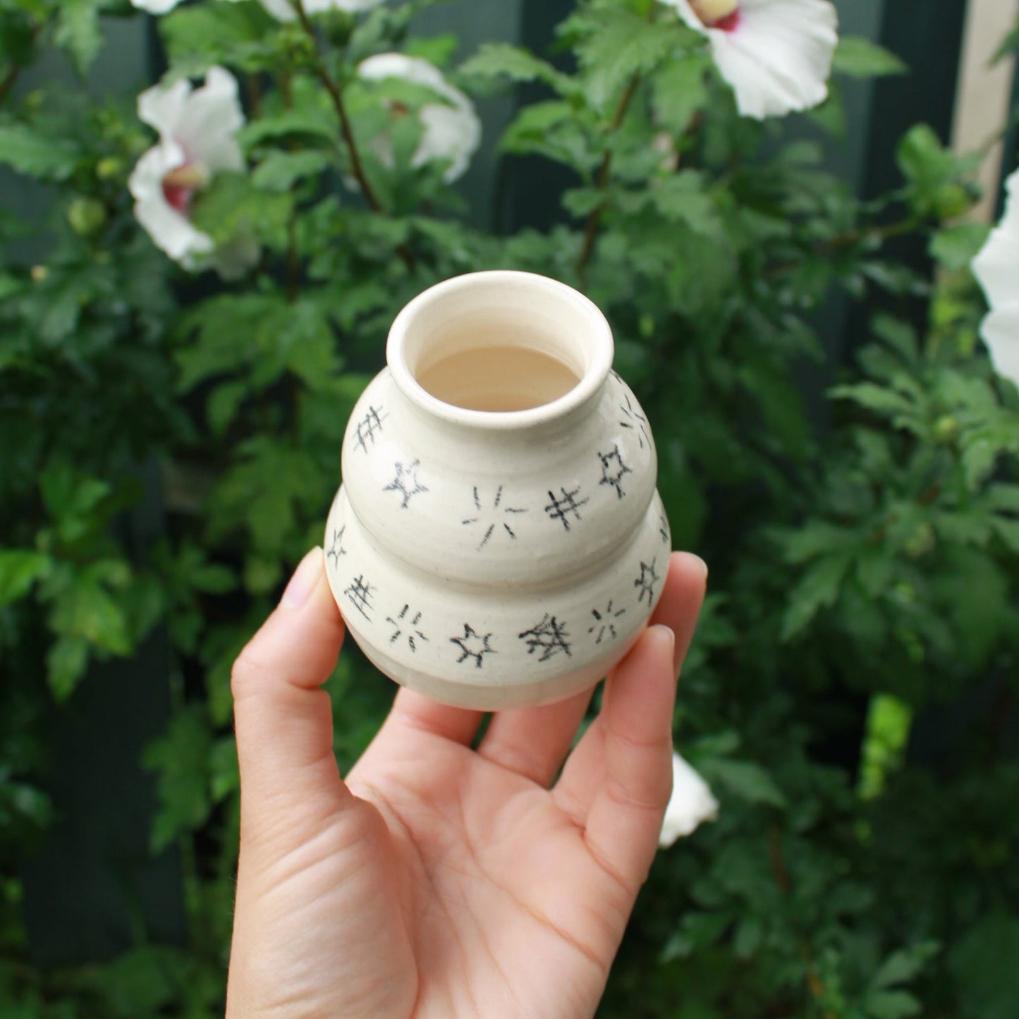 small handthrown bubble vase decorated with hatching and starbursts in black pencil. held in hand against a dark foliage with hibiscus flowers.
