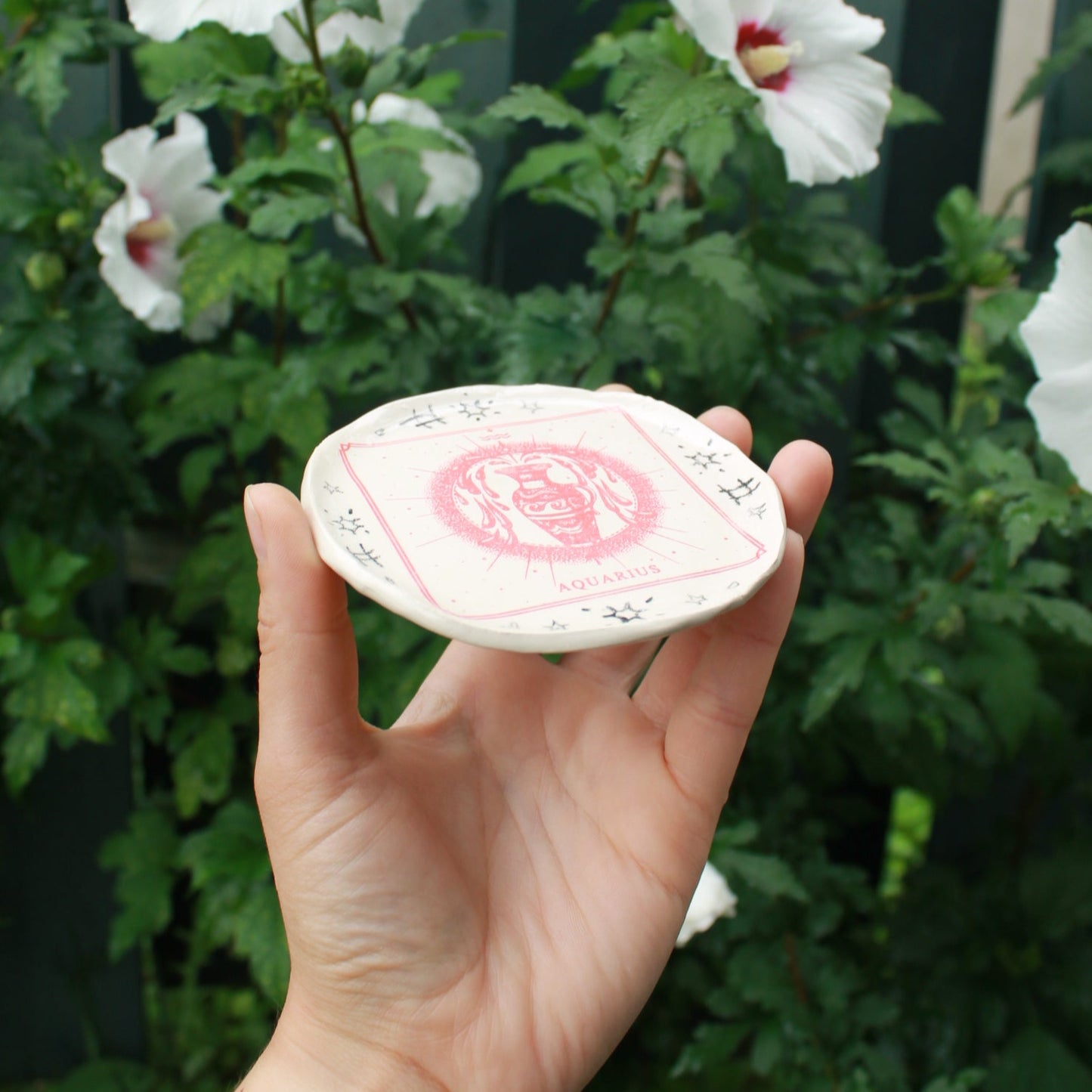 small round jewelry dish decorated with a red square aquarius vase design and surrounded by black pencil doodles. held in hand against a dark foliage background.