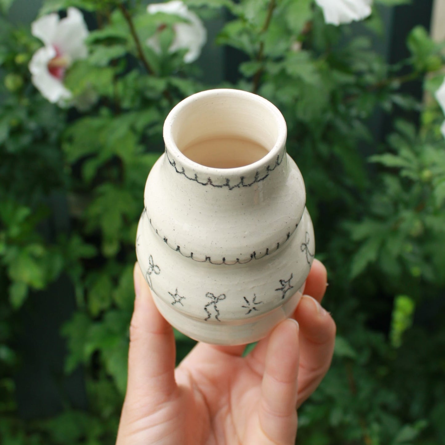 handthrown bubble vase with long neck, decorated with bows, starbursts and scalloped lines. held in hand against a dark foliage background and hibiscus flowers.