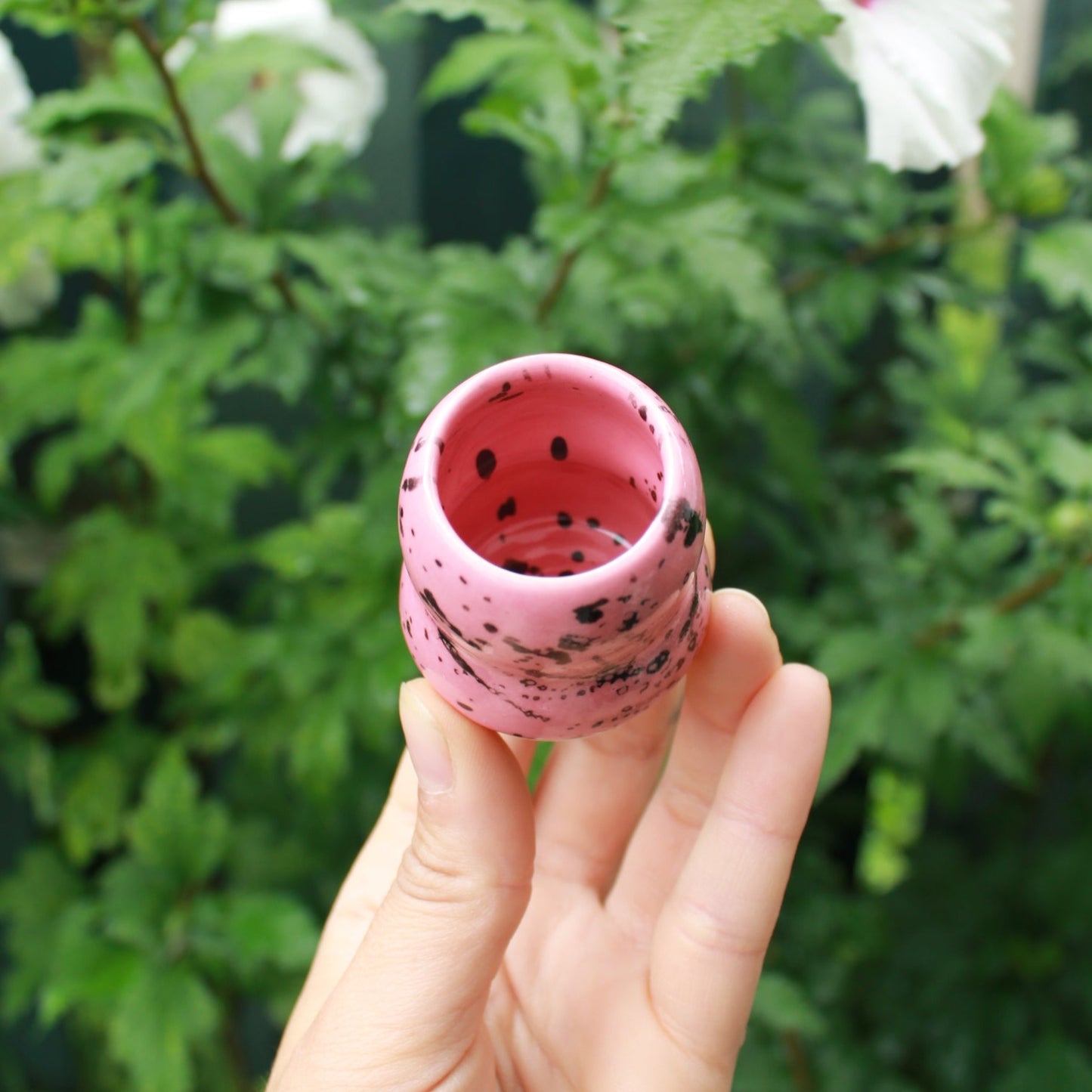 micro bubble bud vase decorated with pink glaze and black splatter. held in hand against a dark foliage background with hibiscus flowers.