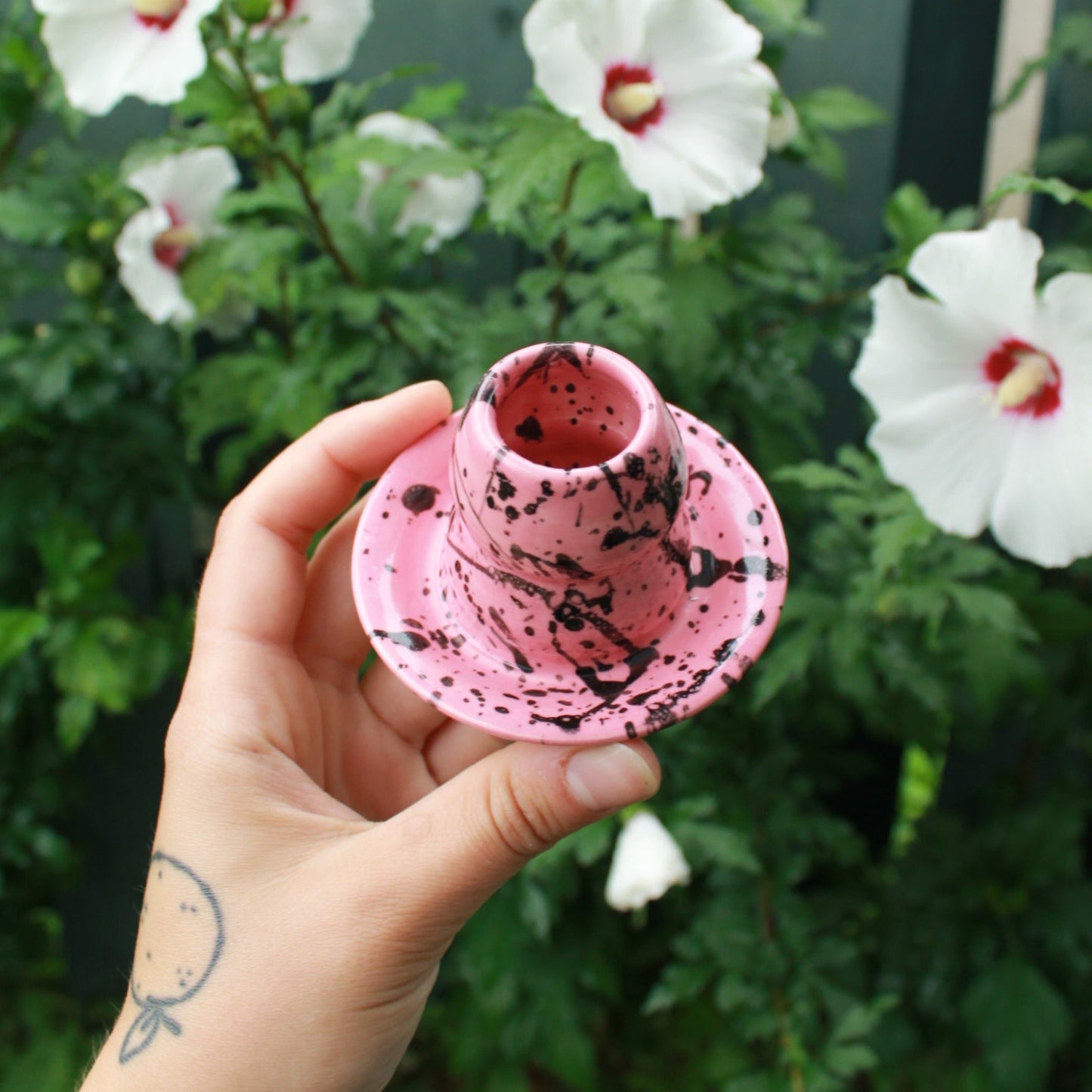 small, curvy, pink candle holder with black splattered glaze. held in hand against a deep green foliage background with white and pink hibiscus flowers peeking out.