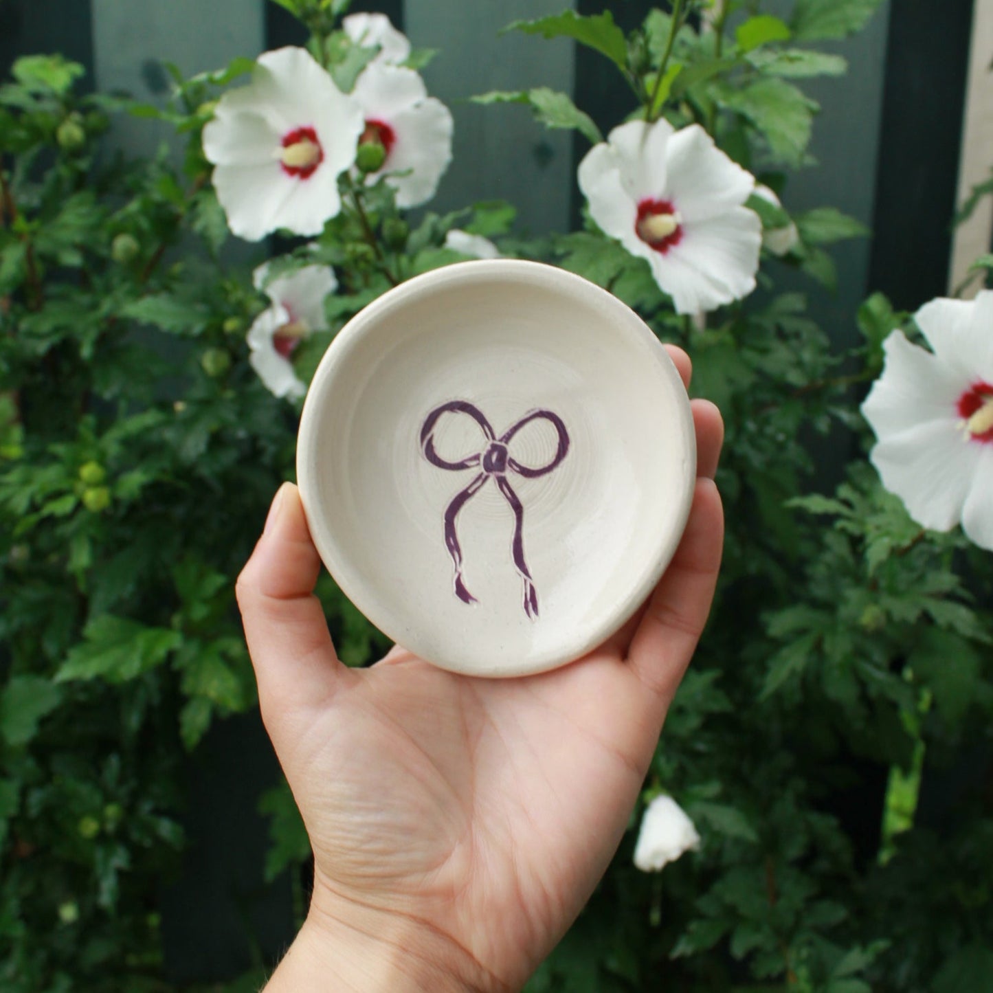 hand thrown off white spoon rest decorated with a dark purple bow in the centre. held in hand against a dark green foliage background, with white and pink hibiscus flowers peeking out.
