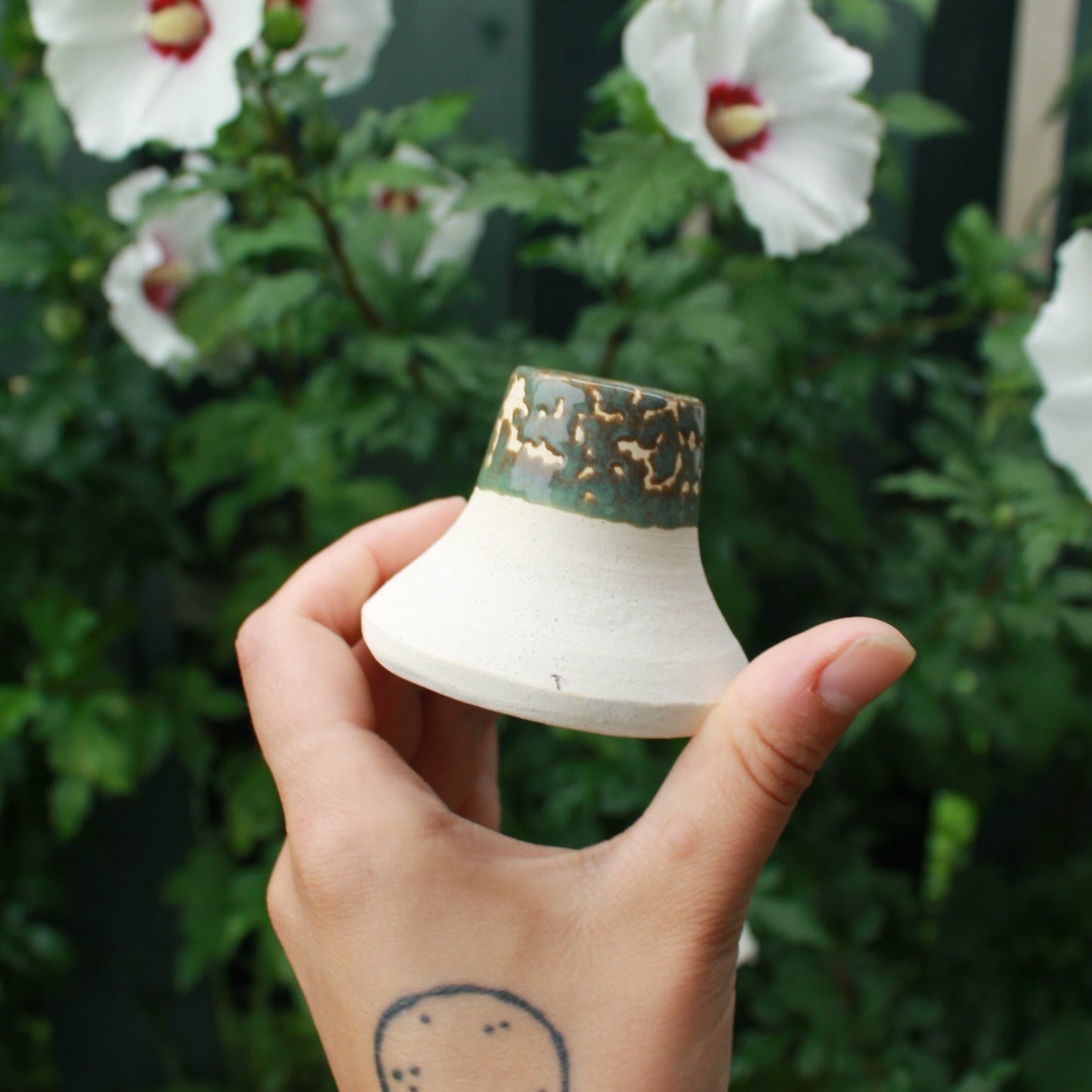 white clay candle holder with crawling sage green glaze. held in hand against a dark foliage background with hibiscus flowers.