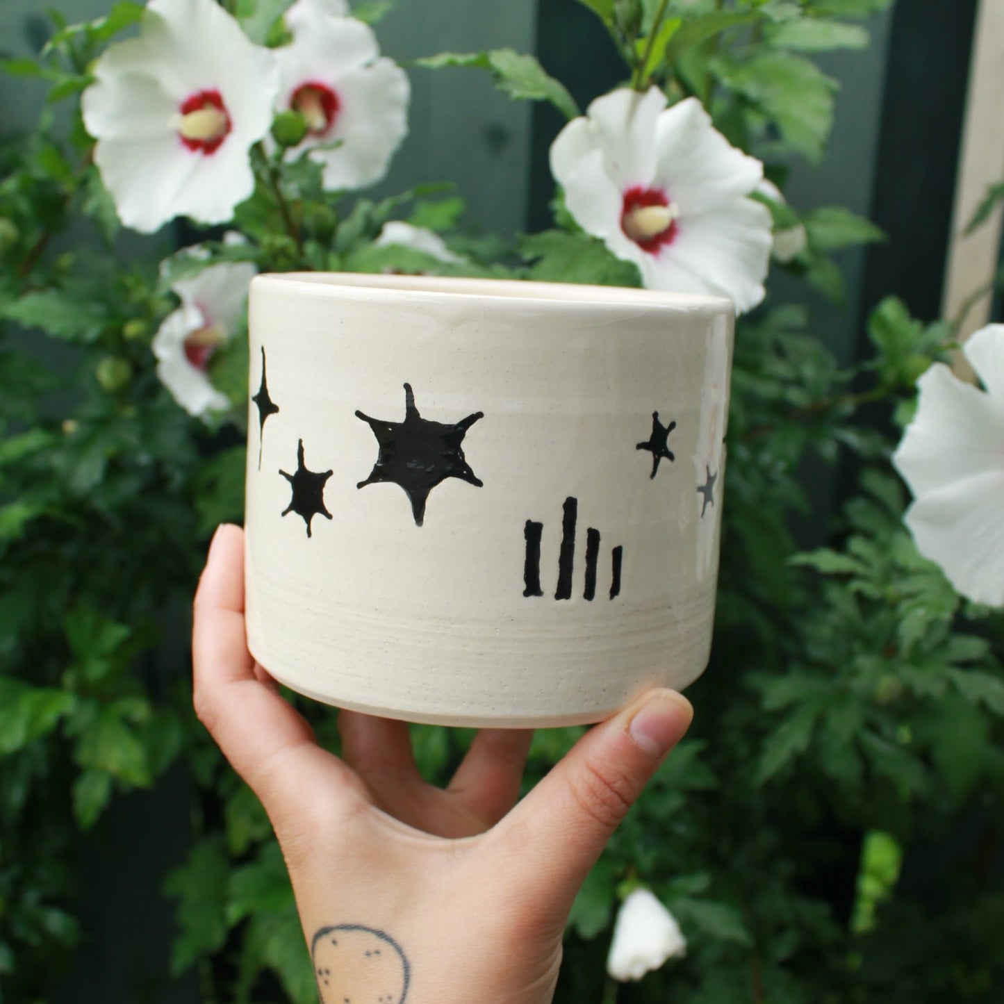large kitchen utensil jug handthrown from white clay and decorated with black glaze celestial designs. held in hand against a dark foliage background with hibiscus flowers.