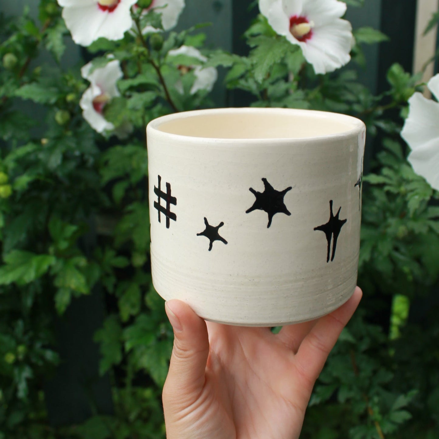 large kitchen utensil jug handthrown from white clay and decorated with black glaze celestial designs. held in hand against a dark foliage background with hibiscus flowers.