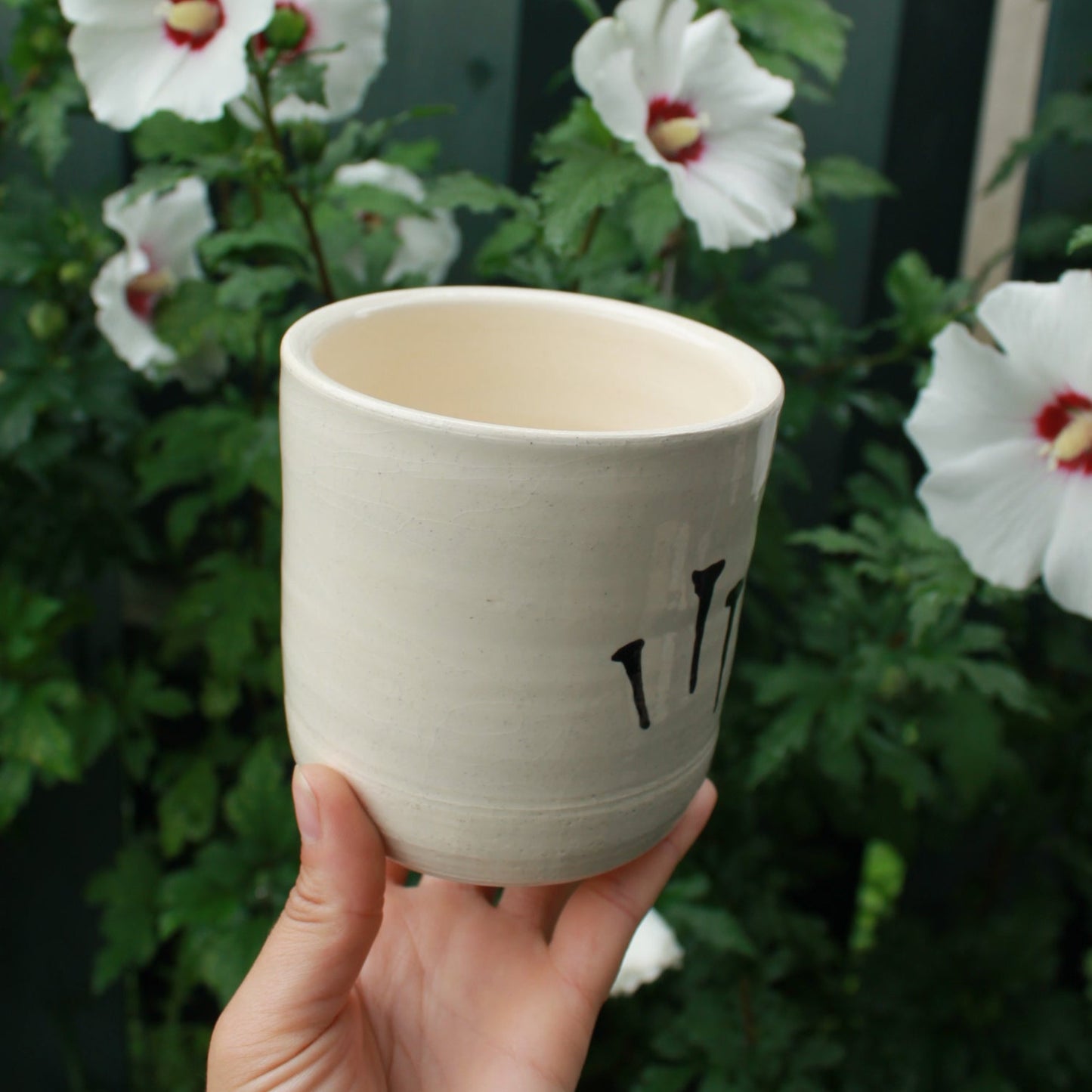 medium sized hand thrown kitchen utensil cup decorated with 5 black glazed nails. held in hand against a dark foliage background with white hibiscus flowers.