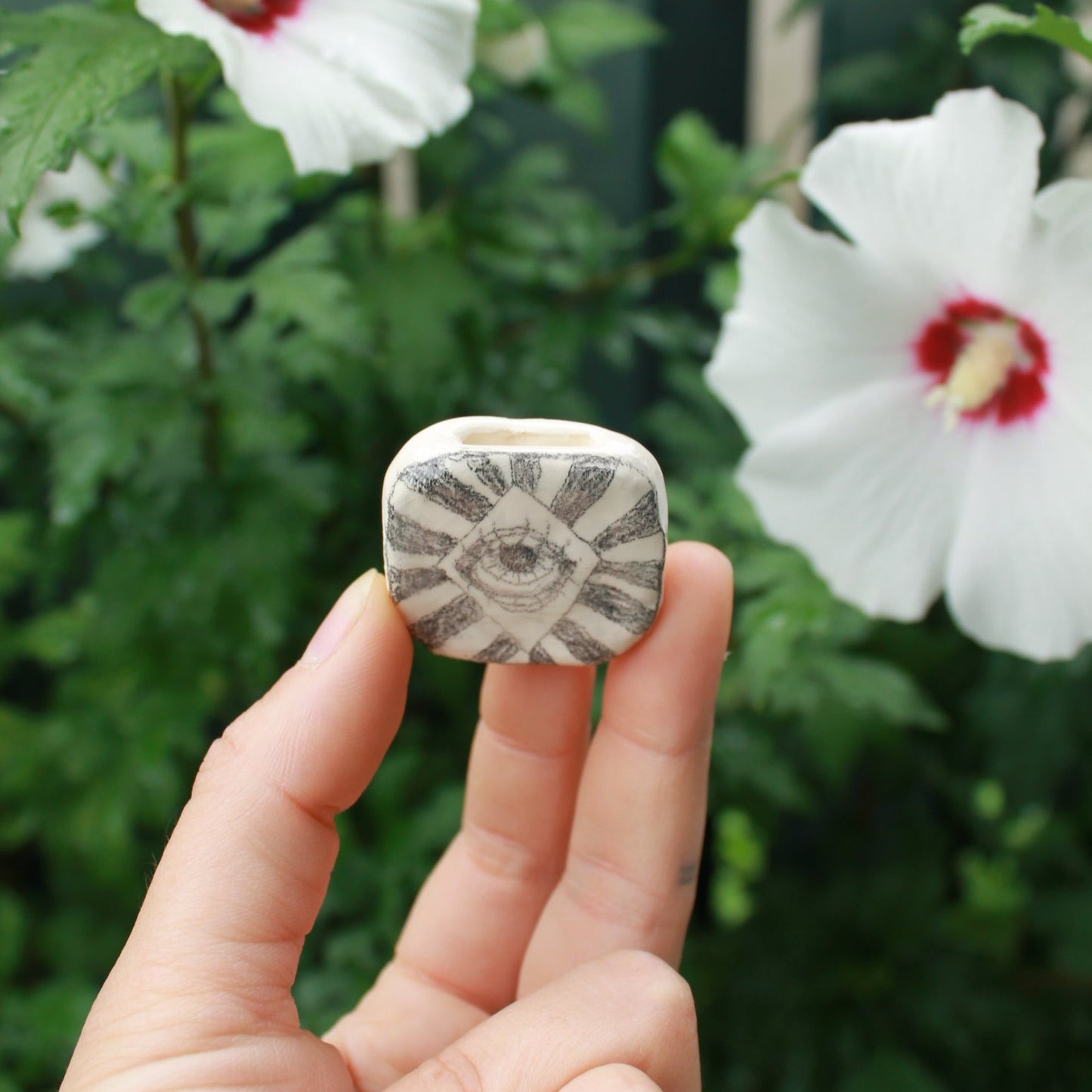 tiny square bud vase decorated with an eye and stripes, held in hand against a dark foliage background and hibiscus flowers.