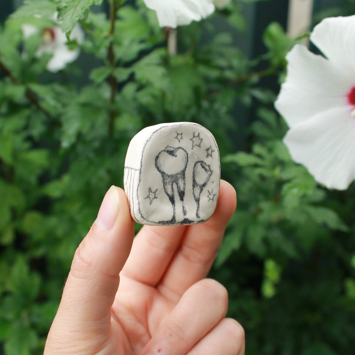 miniature square bud vase decorated with underglaze pencil drawn teeth and starbursts. held in hand against a background of dark foliage and hibiscus flowers.