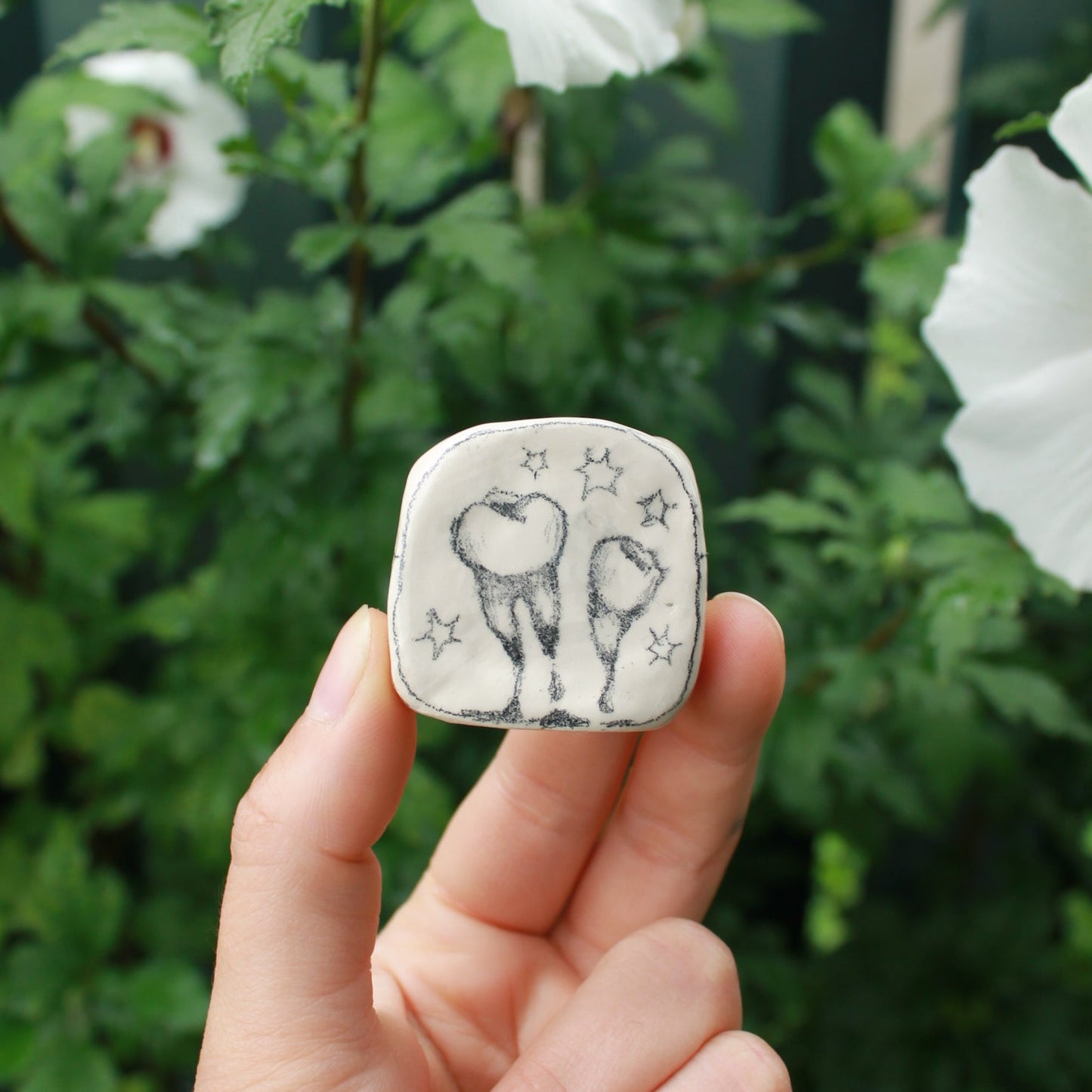 miniature square bud vase decorated with underglaze pencil drawn teeth and starbursts. held in hand against a background of dark foliage and hibiscus flowers.