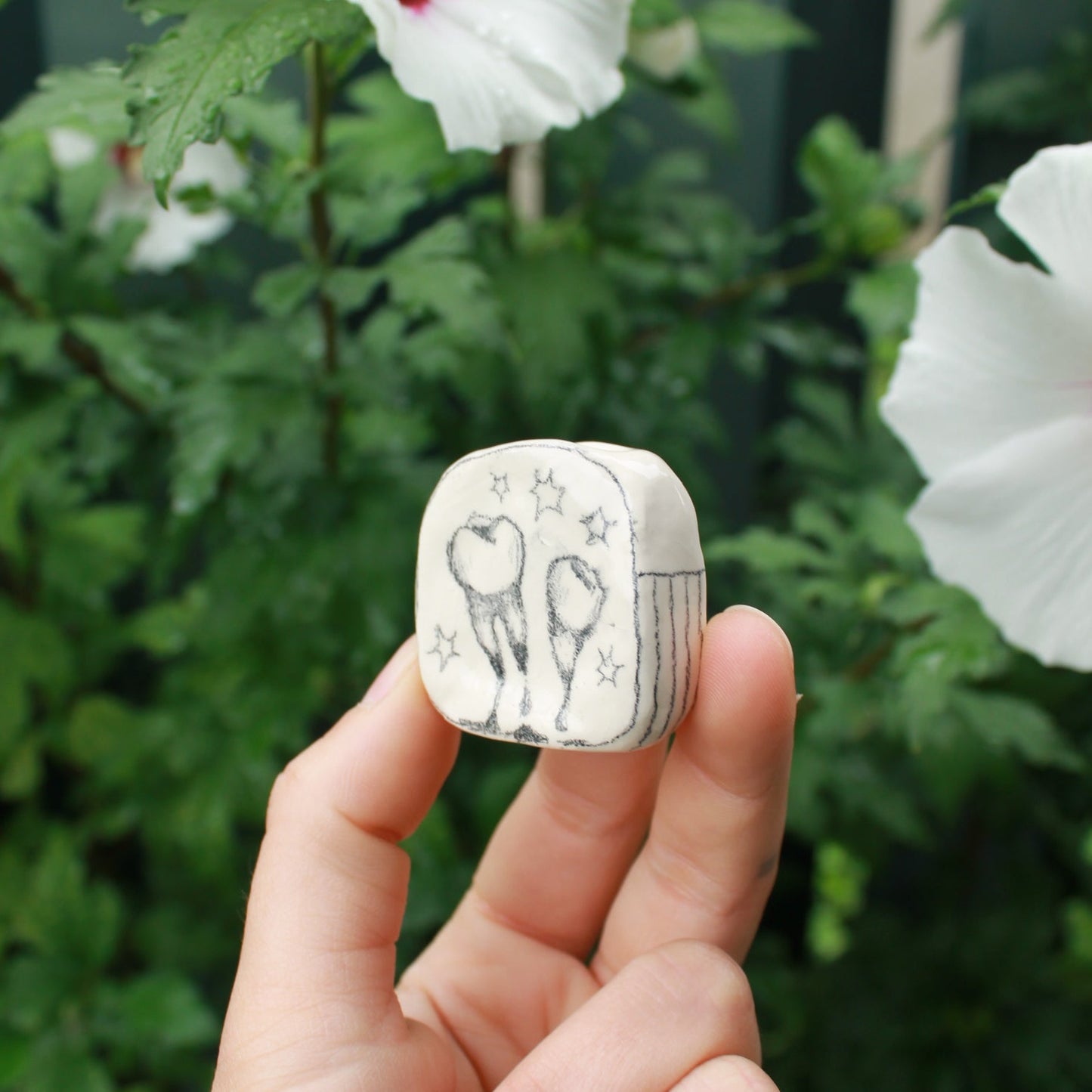 miniature square bud vase decorated with underglaze pencil drawn teeth and starbursts. held in hand against a background of dark foliage and hibiscus flowers.