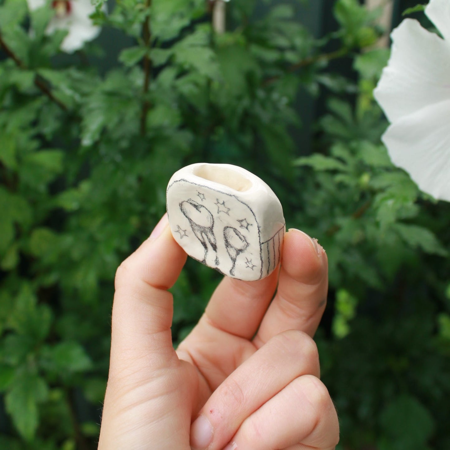 miniature square bud vase decorated with underglaze pencil drawn teeth and starbursts. held in hand against a background of dark foliage and hibiscus flowers.