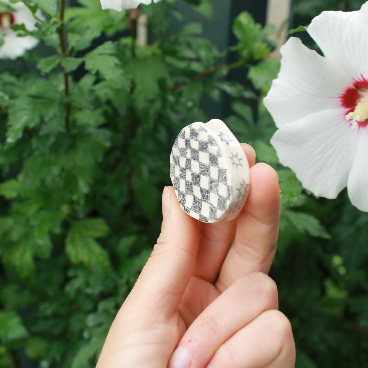 tiny checkered bud vase held in hand against dark foliage with hibiscus flowers.