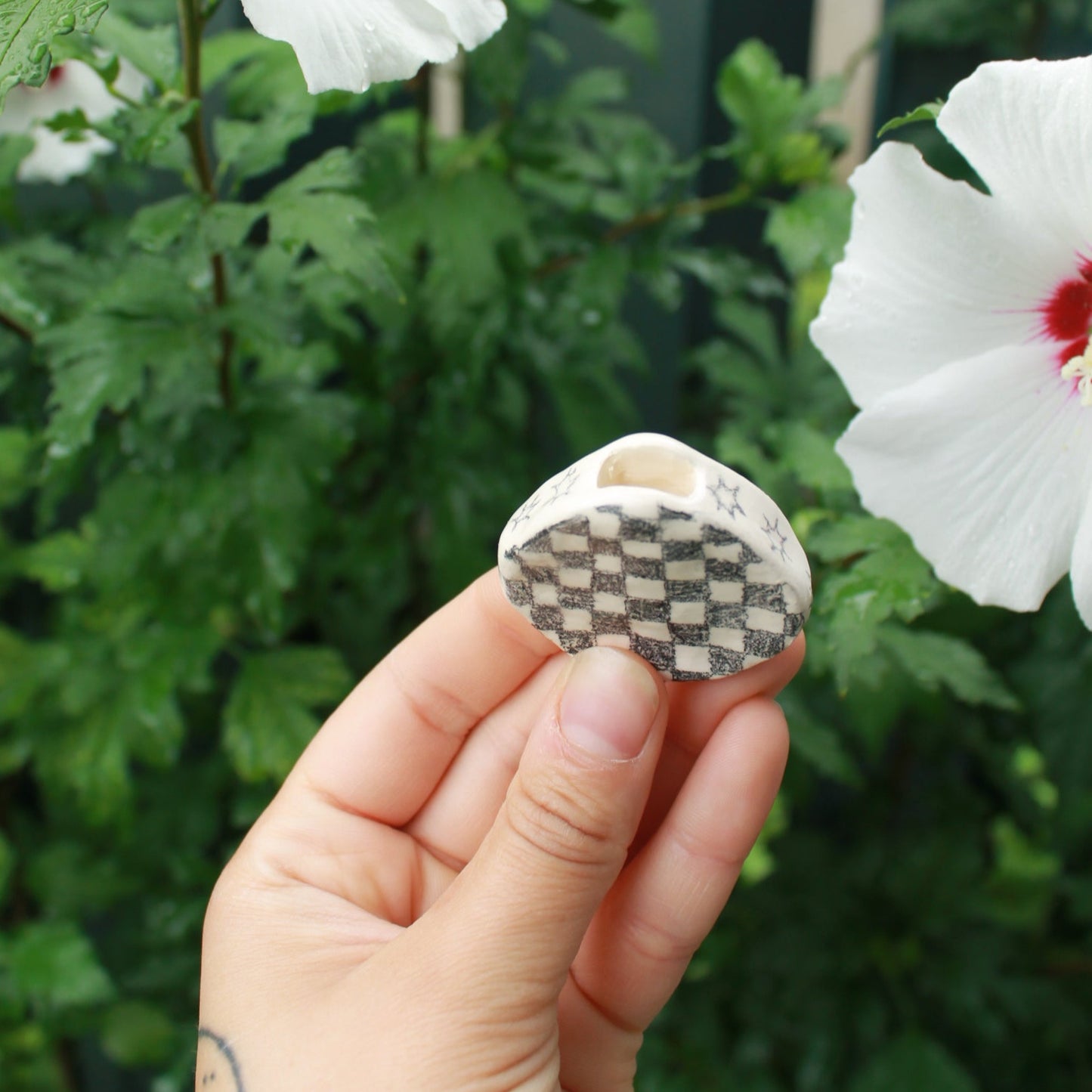 tiny checkered bud vase held in hand against dark foliage with hibiscus flowers.