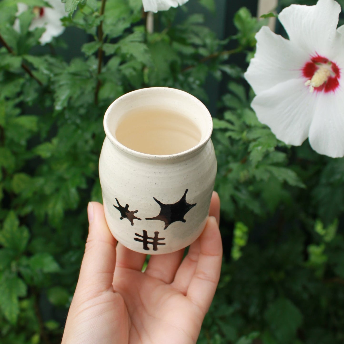 small white jar bud vase decorated with black starburst and hatch designs in the centre. held in hand against a dark foliage backdrop with hibiscus flowers.