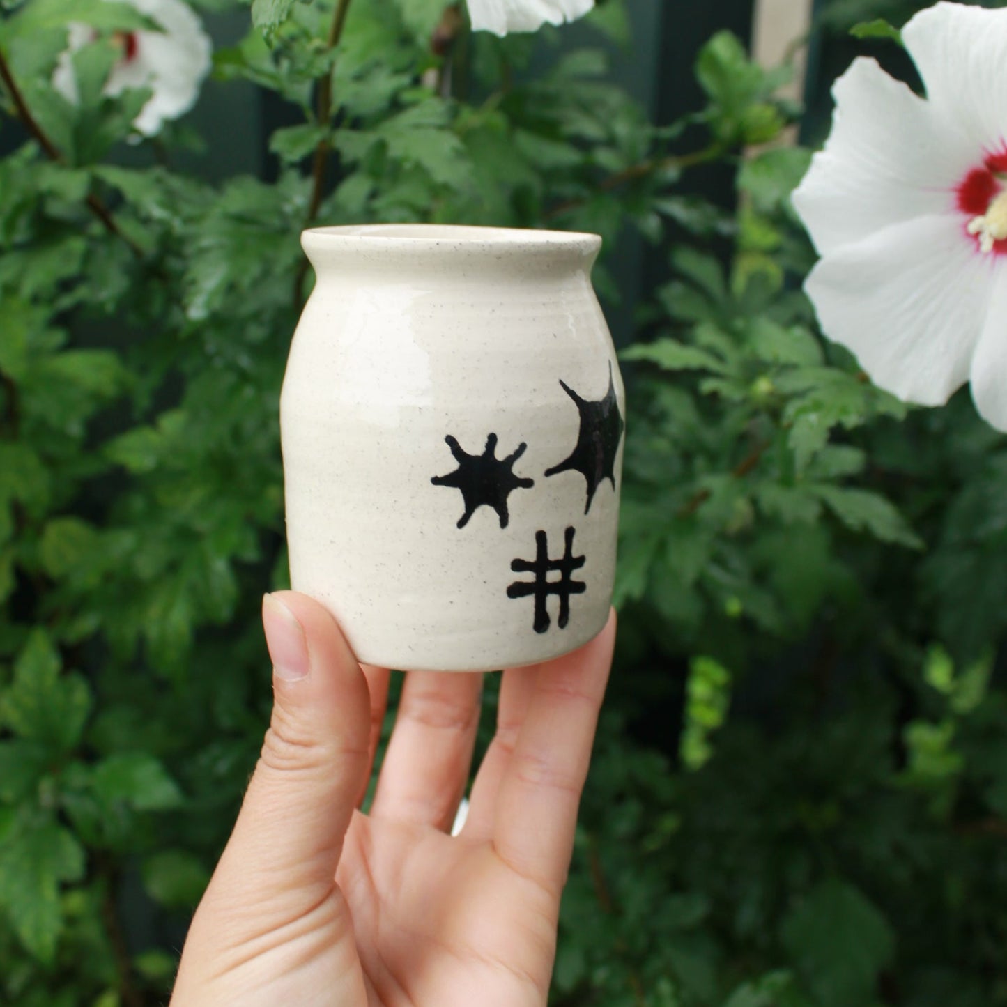 small white jar bud vase decorated with black starburst and hatch designs in the centre. held in hand against a dark foliage backdrop with hibiscus flowers.