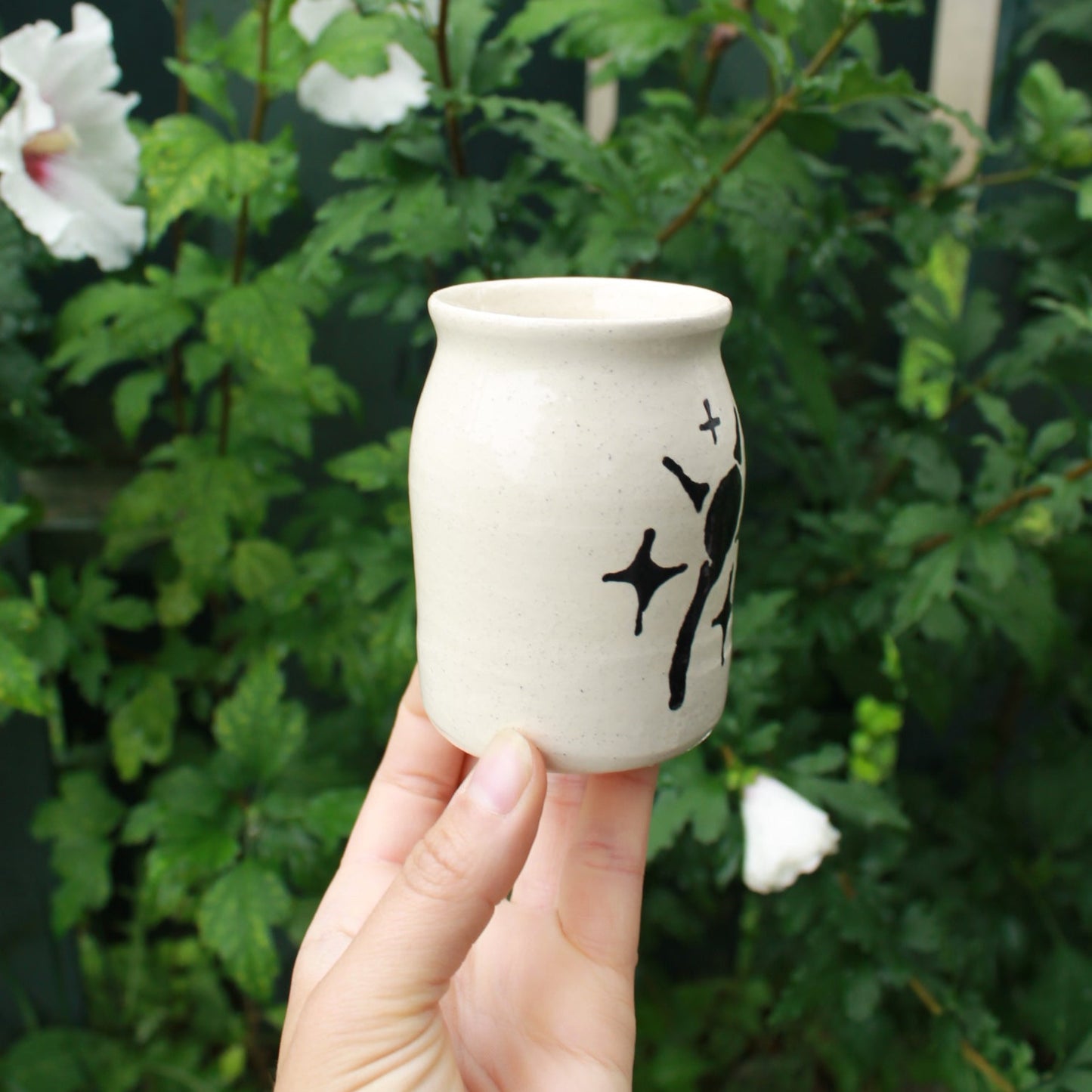 little jar shaped bud vase hand thrown from white clay. decorated with a drippy sun detail and starbursts in black glaze. held in hand against a dark foliage background with white hibiscus flowers.