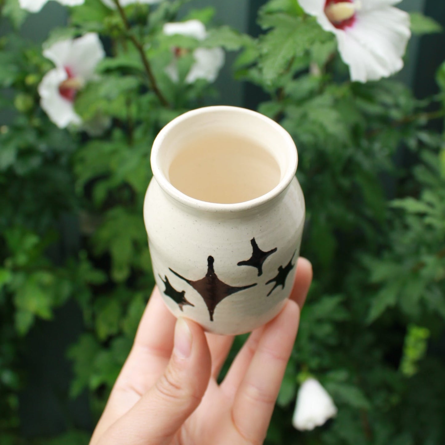 small jar vase made from white clay, decorated with black glaze stars in different shapes and sizes. held in hand against a dark foliage backdrop with white hibiscus flowers.