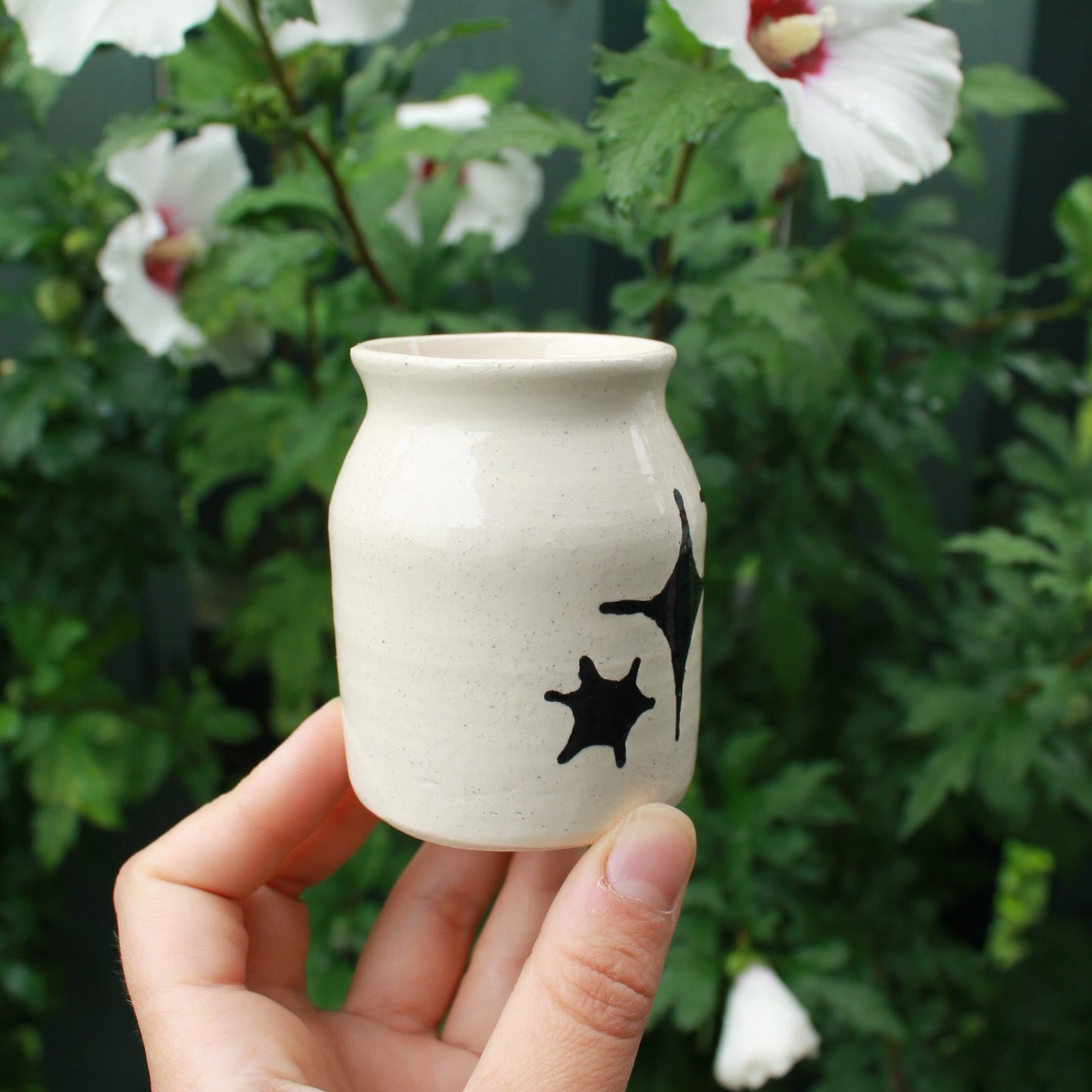 small jar vase made from white clay, decorated with black glaze stars in different shapes and sizes. held in hand against a dark foliage backdrop with white hibiscus flowers.