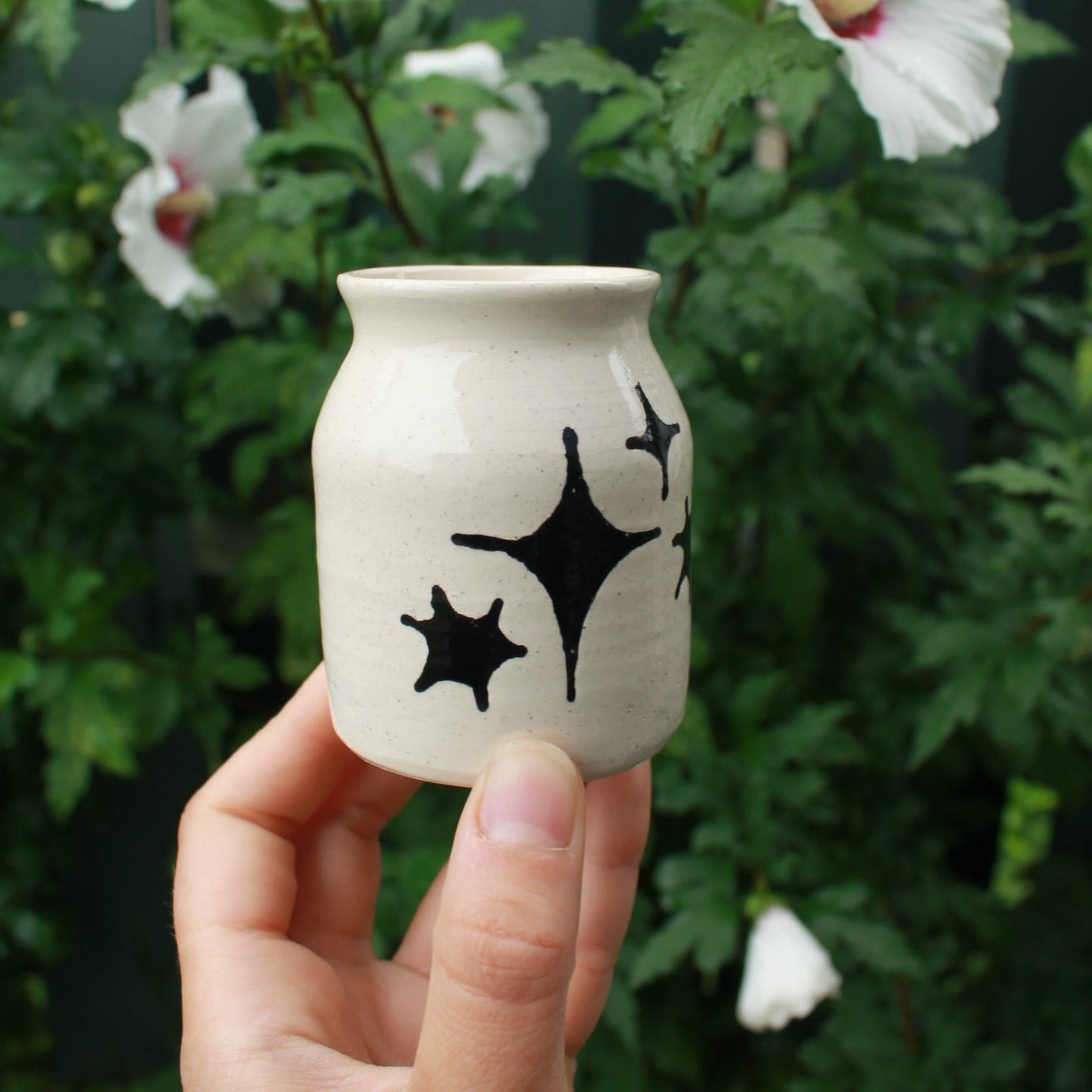 small jar vase made from white clay, decorated with black glaze stars in different shapes and sizes. held in hand against a dark foliage backdrop with white hibiscus flowers.
