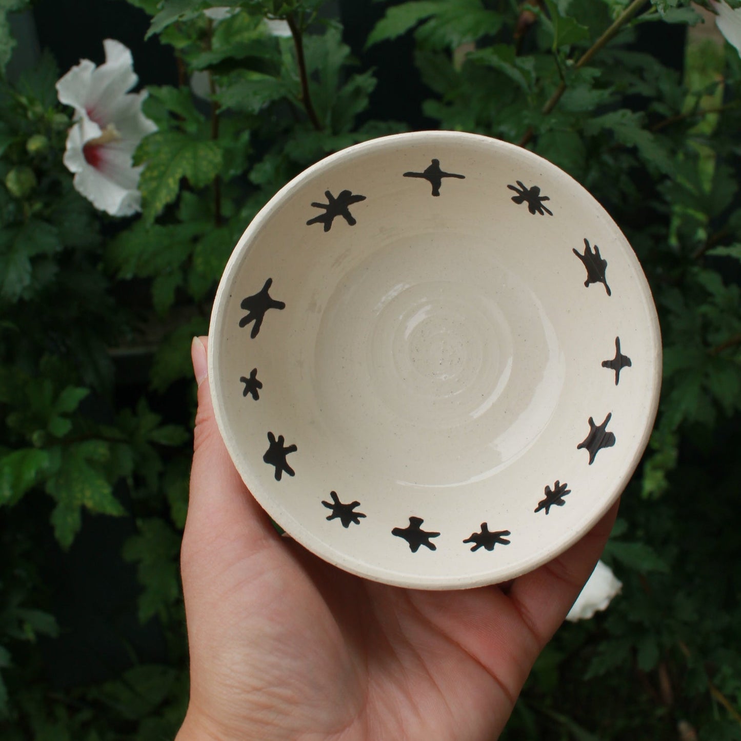 small white bowl with black stars decorating the inside rim. held in hand against a dark foliage background with hibiscus flowers.