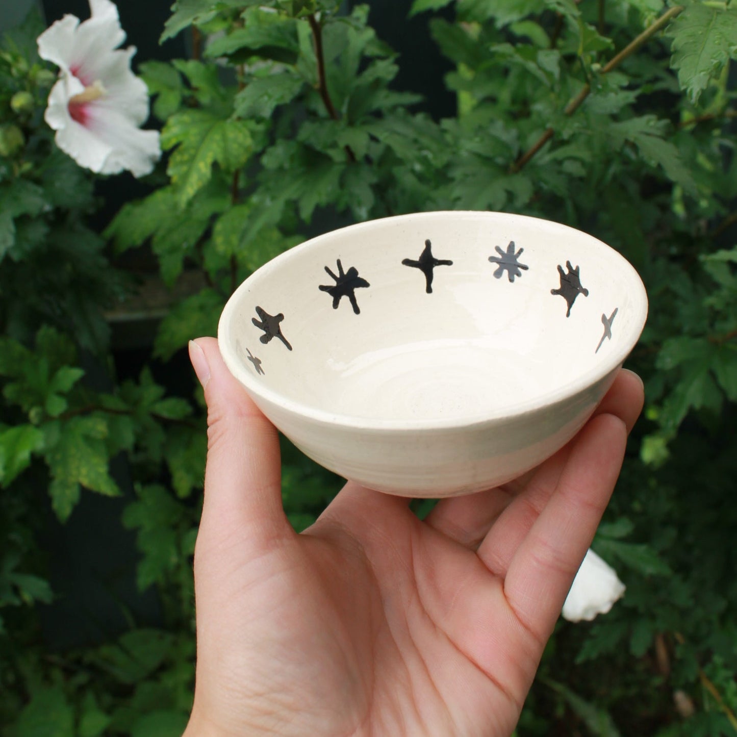 small white bowl with black stars decorating the inside rim. held in hand against a dark foliage background with hibiscus flowers.