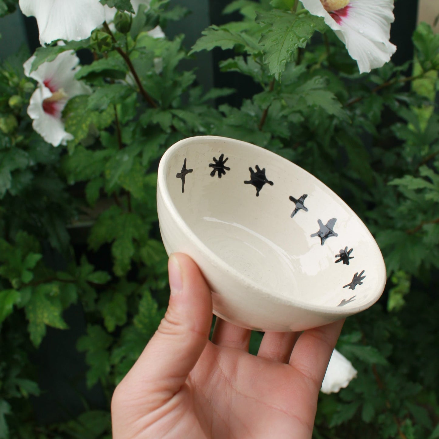 small white bowl with black stars decorating the inside rim. held in hand against a dark foliage background with hibiscus flowers.