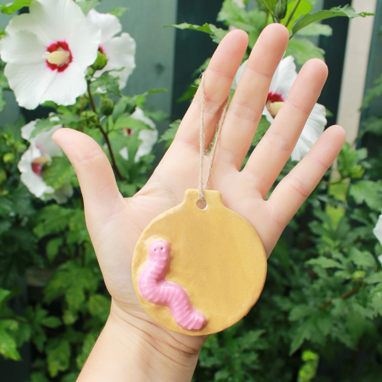 flat circular ornaments with pink worms appliqued to them. the design of this one is a light brown dirt worm. held in hand against a dark green foliage background, with white and pink hibiscus flowers peeking through. 