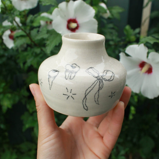 Hand holding a small ceramic vase with a flowy bow and teeth designs drawn on it. In the background are some white hardy hibiscus flowers tucked into some dark green foliage.