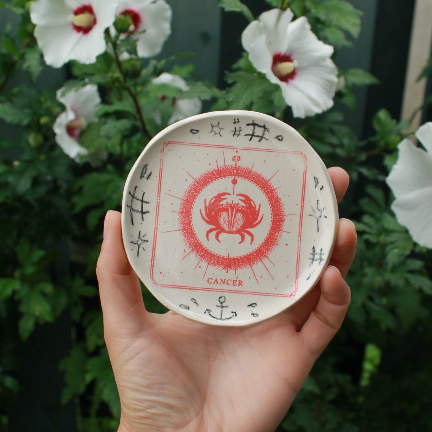 small round jewelry dish decorated with a red square cancer crab design. black pencil doodles surround it. held in hand on an angle against a dark foliage background with hibiscus flowers. 