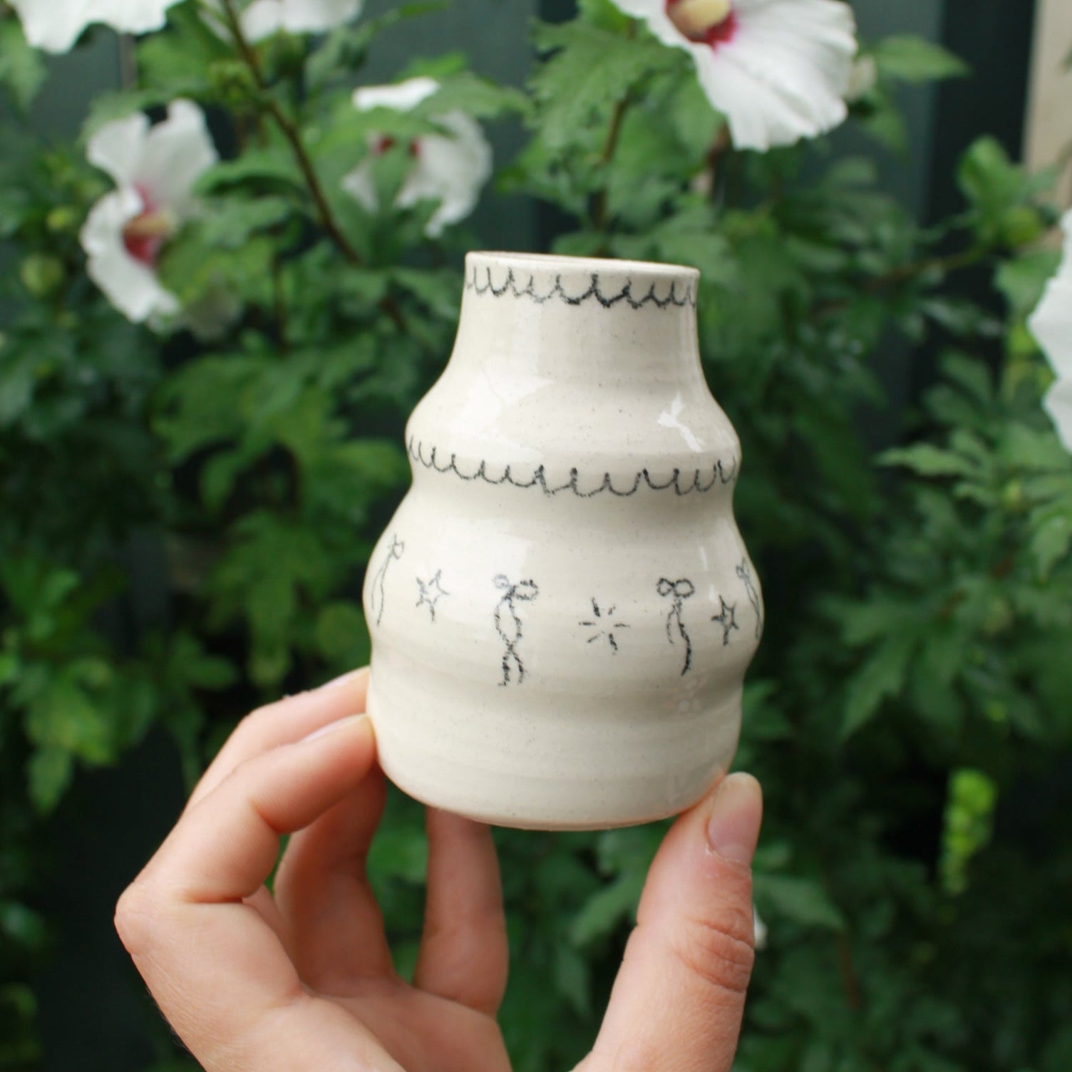 handthrown bubble vase with long neck, decorated with bows, starbursts and scalloped lines. held in hand against a dark foliage background and hibiscus flowers.