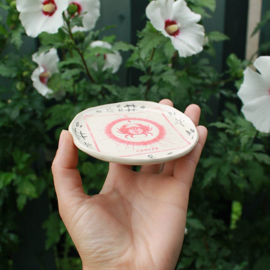 small round jewelry dish decorated with a red square cancer crab design. black pencil doodles surround it. held in hand on an angle against a dark foliage background with hibiscus flowers. 