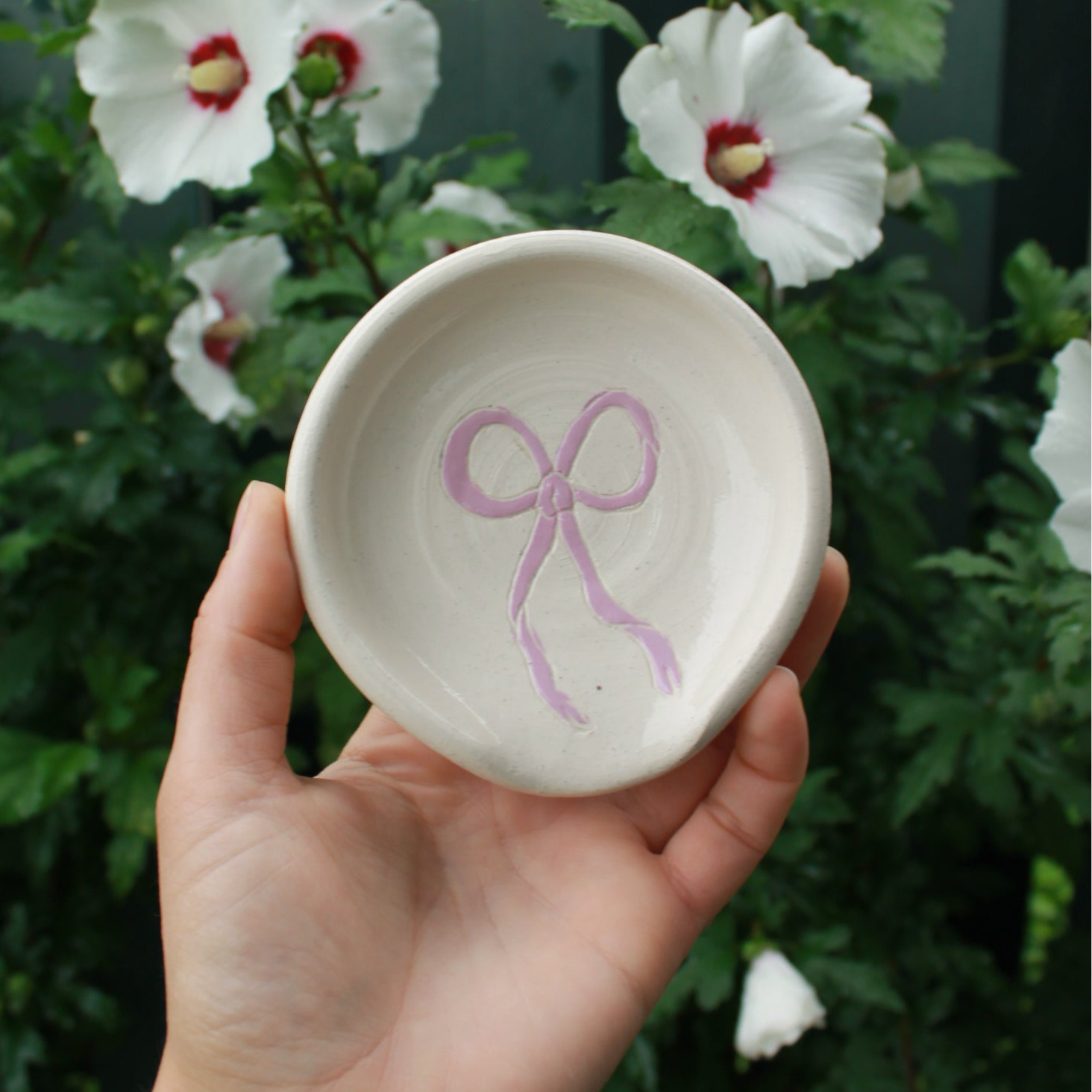 hand thrown off white spoon rests decorated with a lavender purple bow in the centre. held in hand against a dark green foliage background, with white and pink hibiscus flowers peeking out. 
