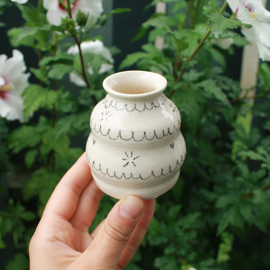 handthrown bubbly bud vase made from white clay, decorated with black pencil scalloped lines and tiny starbursts and hearts. held in hand against a dark foliage background with white hibiscus flowers. 