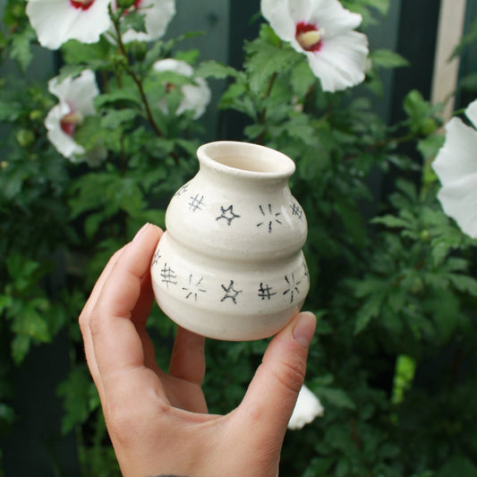 small handthrown bubble vase decorated with hatching and starbursts in black pencil. held in hand against a dark foliage with hibiscus flowers. 