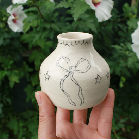hand holding a small off white ceramic vase with star and bow design, and a scalloped edge drawn around the opening. background is dark green foliage with pink and white hibiscus flowers peeking out.