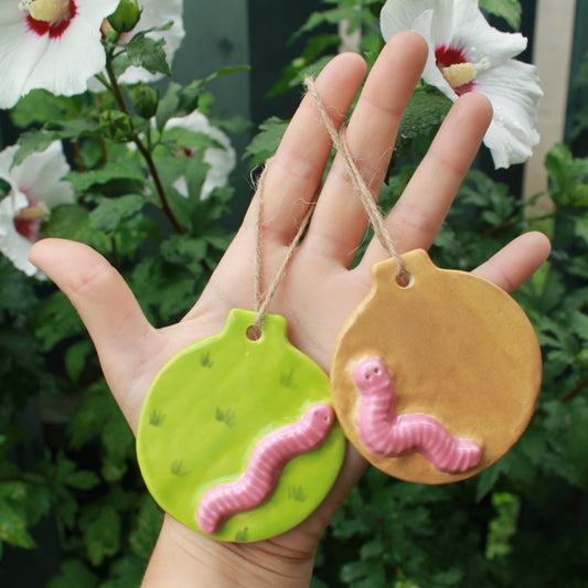 flat circular ornaments with pink worms appliqued to them. One is a green meadow worm, and the other is a light brown dirt worm. held in hand against a dark green foliage background, with white and pink hibiscus flowers peeking through. 