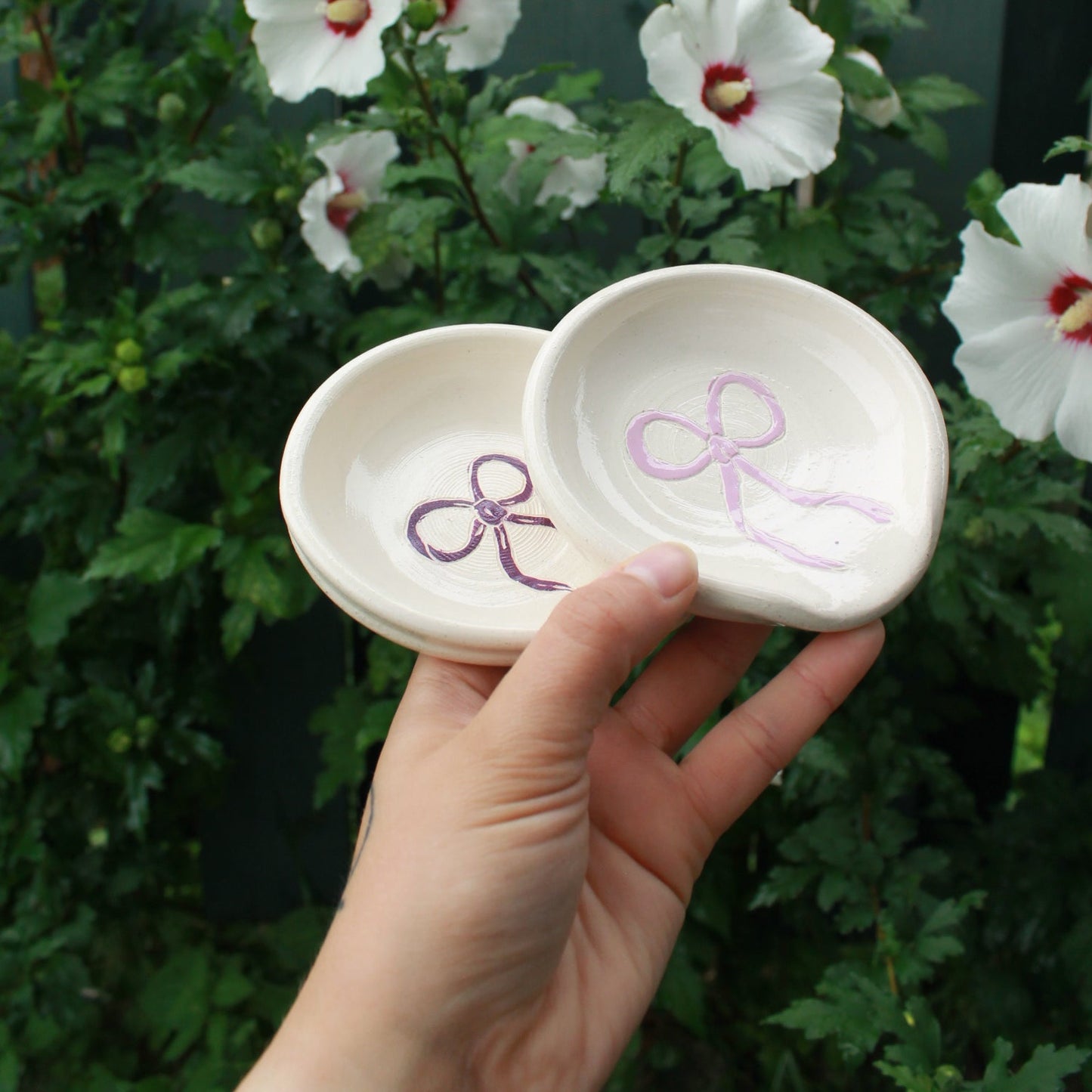 hand thrown off white spoon rests decorated with a purple bow in the centre. held in hand against a dark green foliage background, with white and pink hibiscus flowers peeking out. 